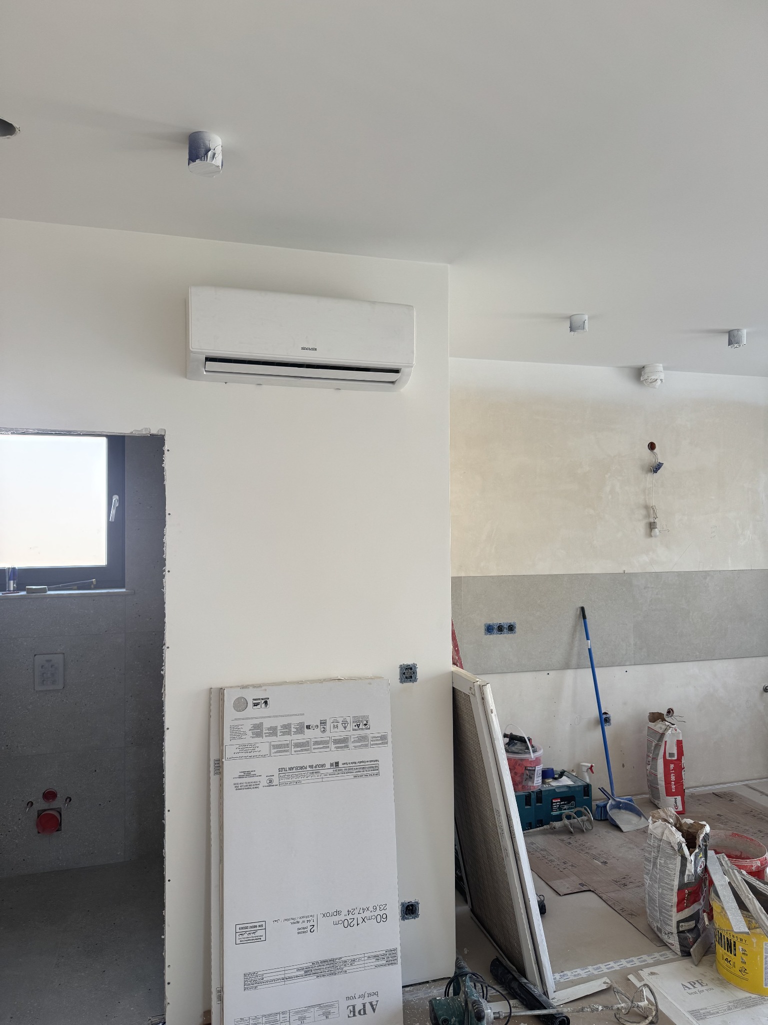 Biała klimatyzacja Sinclair na ścianie w surowym wnętrzu. Widoczne materiały budowlane, płytki i narzędzia. W tle ściana w trakcie wykończenia.