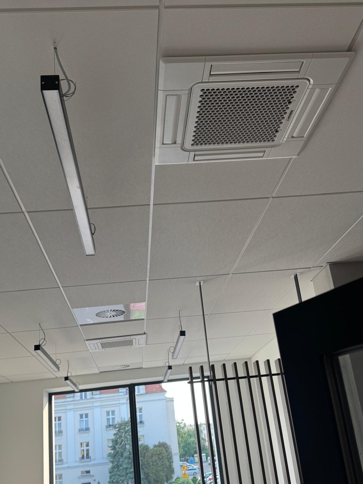 Sufit biurowy z zamontowaną kasetonową klimatyzacją i oświetleniem liniowym LED. Widok z dołu na system wentylacyjny i okno z widokiem na budynek.
