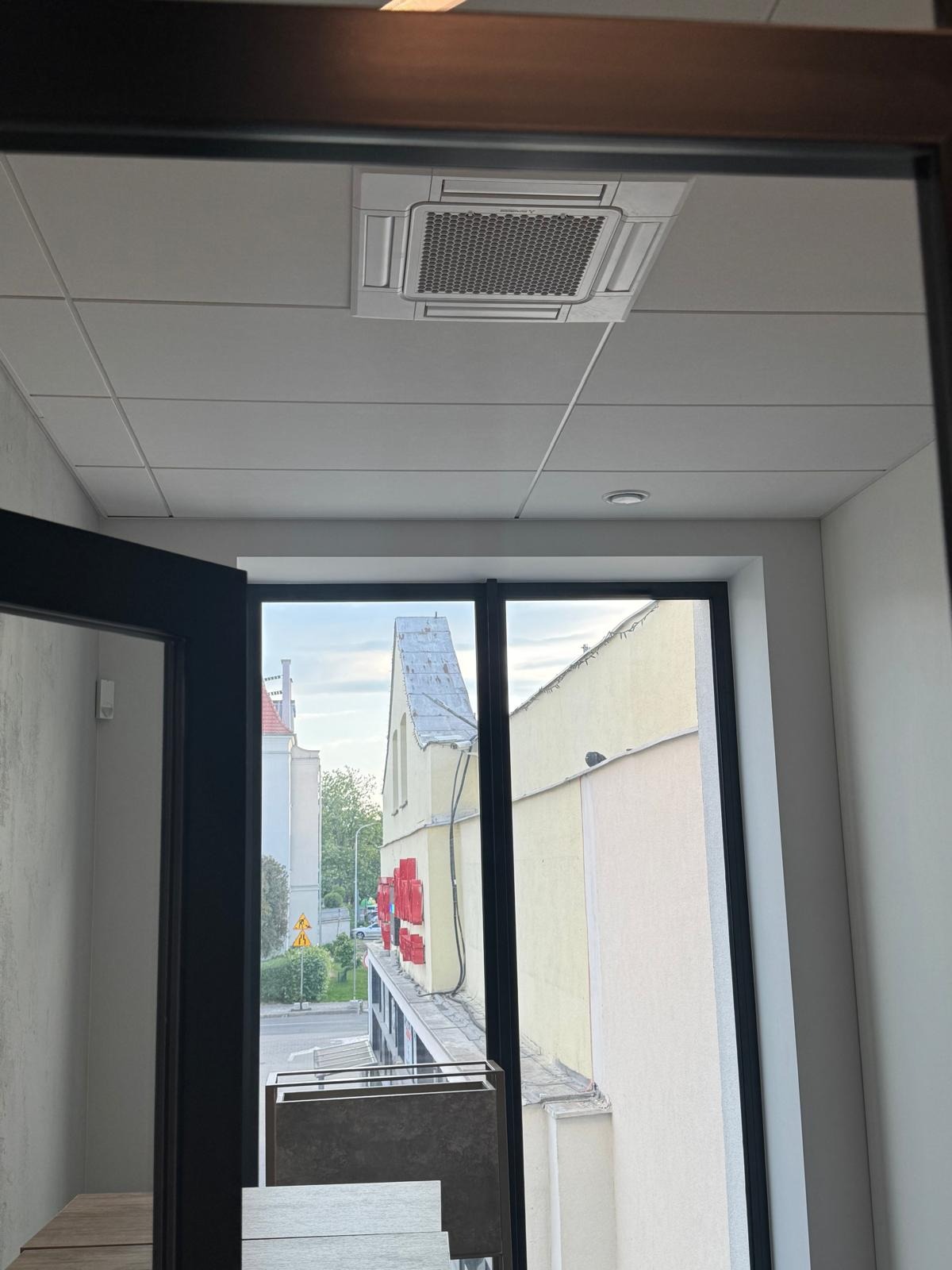 Kasetonowa klimatyzacja sufitowa w biurze w Sady. Widok przez okno na budynek z czerwonymi elementami dekoracyjnymi. Nowoczesny system klimatyzacji.