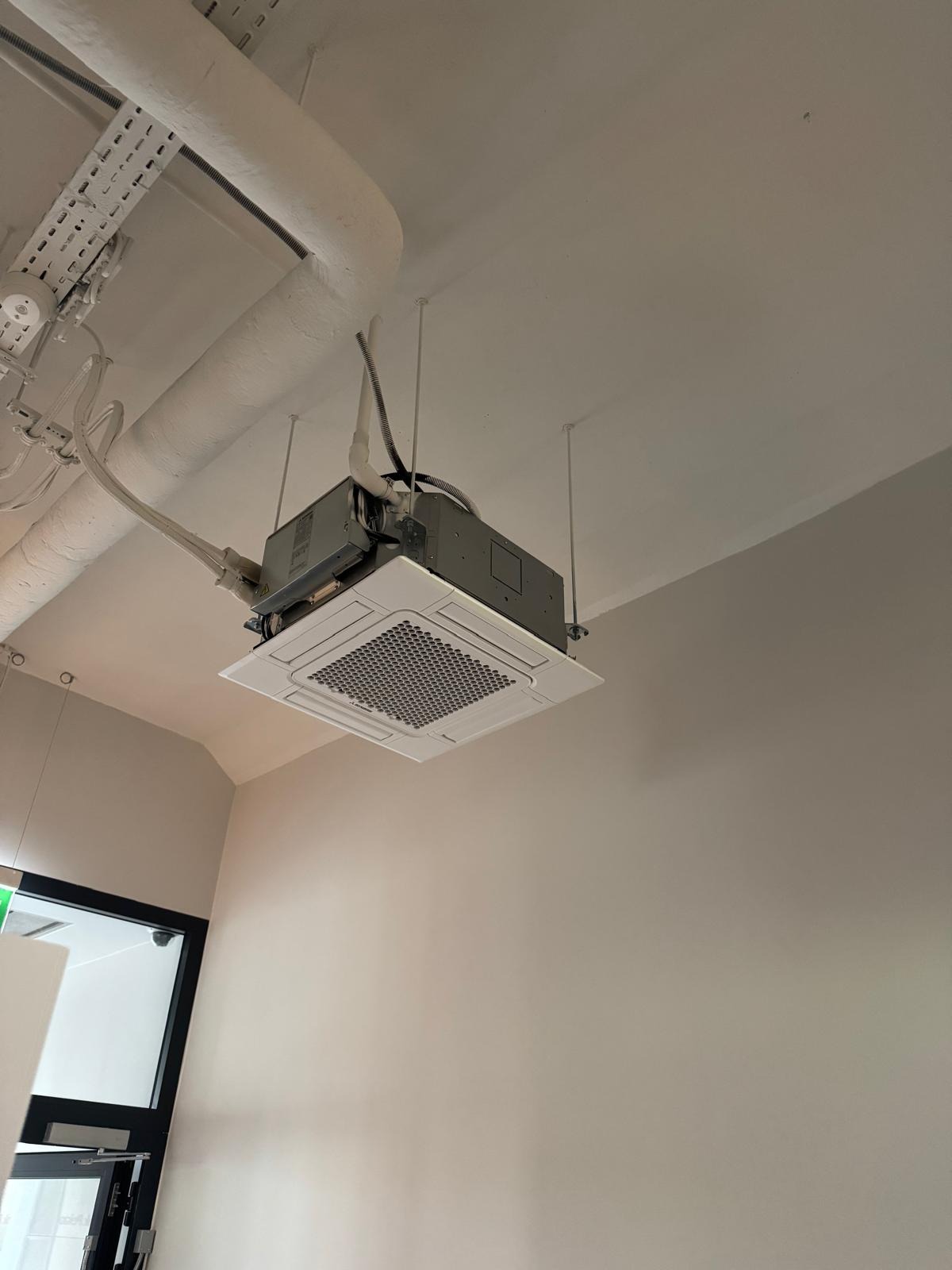 Sufitowa klimatyzacja kasetonowa z widocznymi przewodami i instalacją, zamontowana w jasnym pomieszczeniu biurowym. Widok z dołu.
