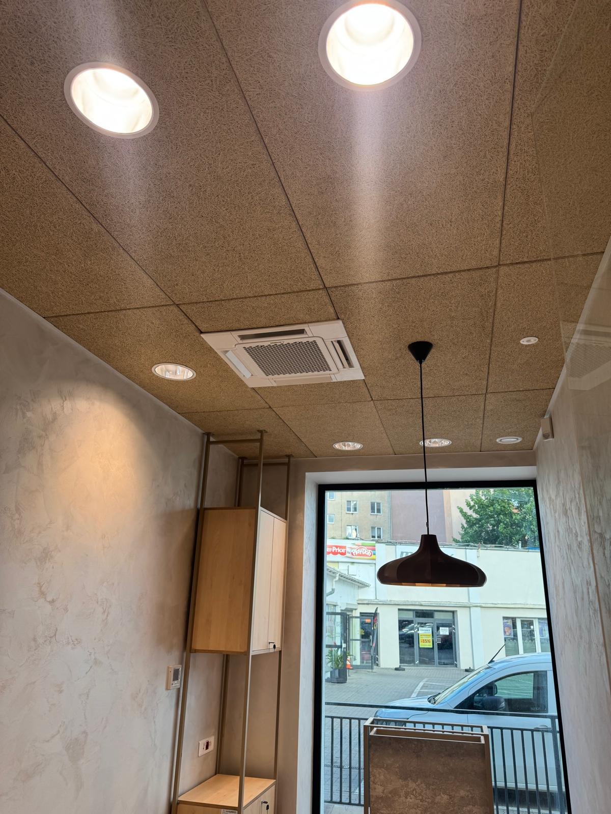Kasetonowa struktura sufitu z wbudowaną klimatyzacją kasetonową, oświetleniem punktowym i wiszącą lampą. Widok z dołu na wnętrze lokalu.