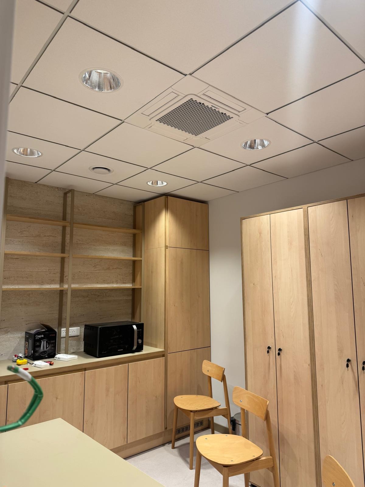 Kasetonowa klimatyzacja sufitowa w jasnym pomieszczeniu biurowym z drewnianymi szafkami i oświetleniem punktowym. Widok na zamontowane urządzenie.