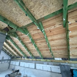 SoriAn - Skośny dach poddasza z drewnianą konstrukcją, zielonymi belkami i metalowymi łącznikami. Widoczne płyty OSB i fragment muru z betonu komórkowego.