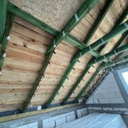 SoriAn - Drewniana konstrukcja dachu poddasza z zielonymi belkami, widoczne mocowania i folia aluminiowa, przygotowana do ocieplenia. Ujęcie z dołu, widoczne okno dachowe.