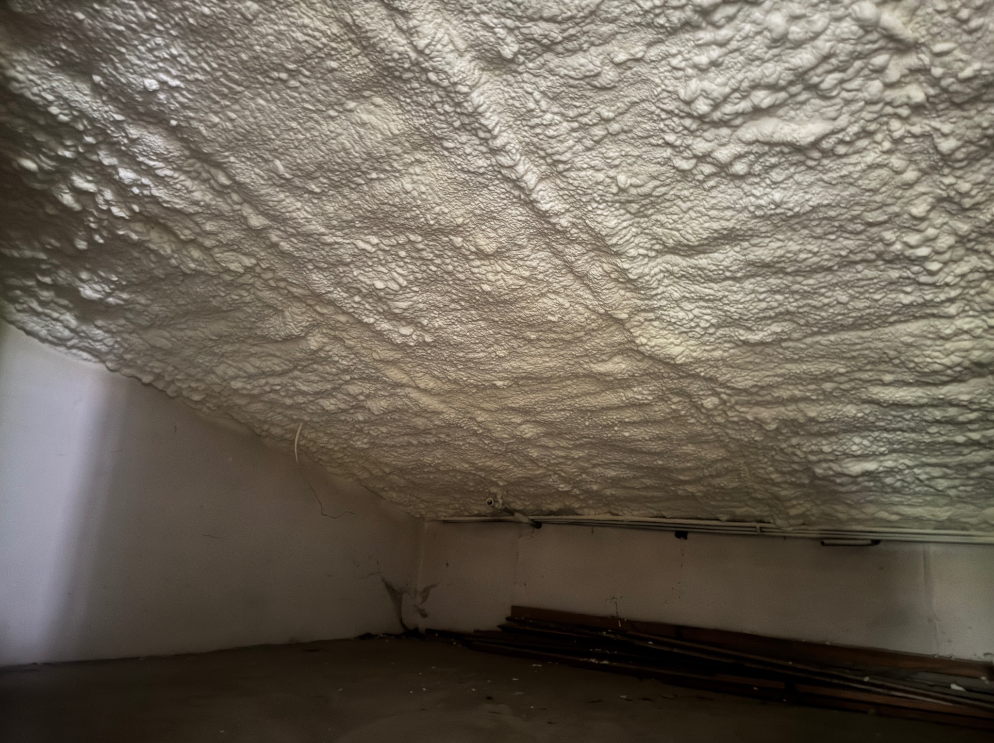 Strych po ociepleniu pianką PUR. Widoczne białe, nieregularne struktury pianki pokrywające skośny sufit i fragment ściany. Elementy instalacji pod sufitem.
