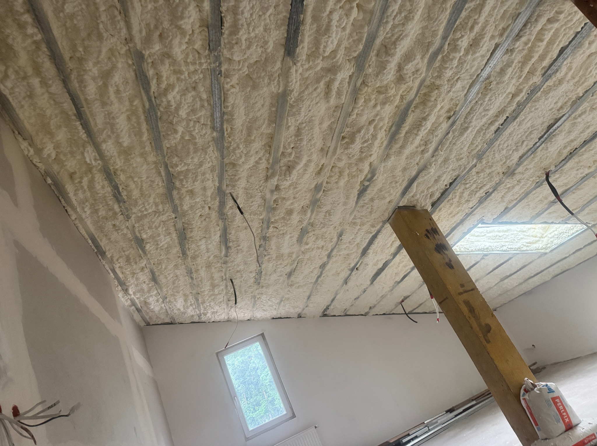 Poddasze z widoczną izolacją pianką PUR między metalowymi profilami, okno dachowe w tle, surowy stan wykończenia wnętrza.