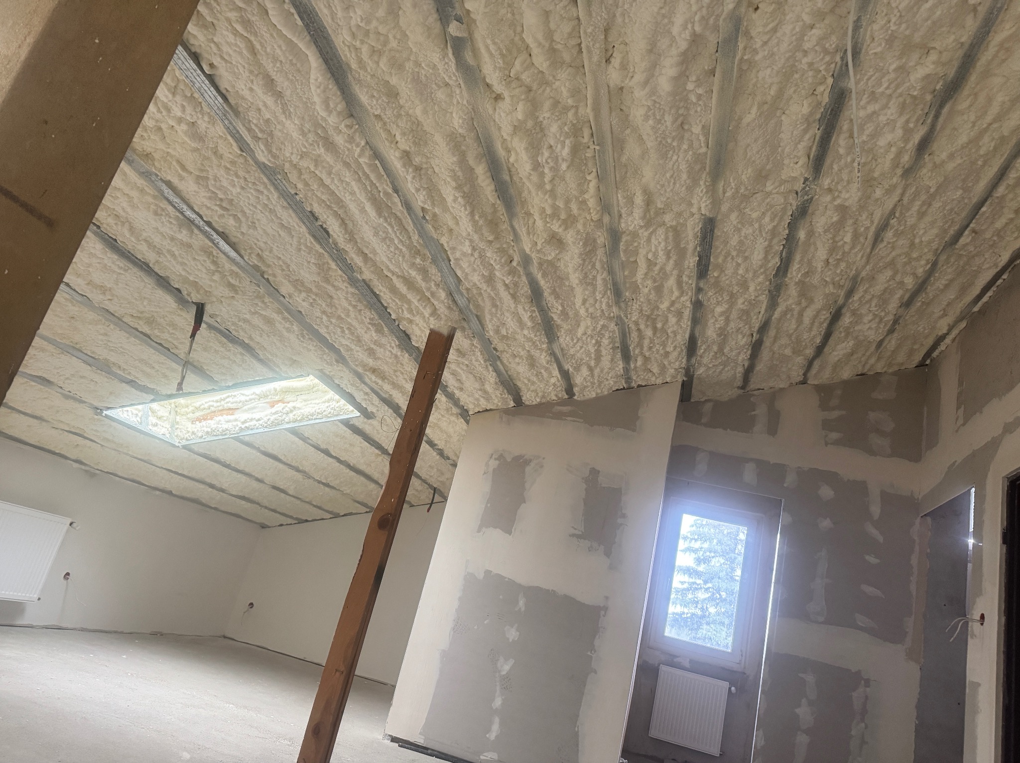 Poddasze po ociepleniu pianką PUR, widoczne metalowe profile i równomiernie nałożona pianka. W tle okno i fragment ściany w trakcie prac wykończeniowych.