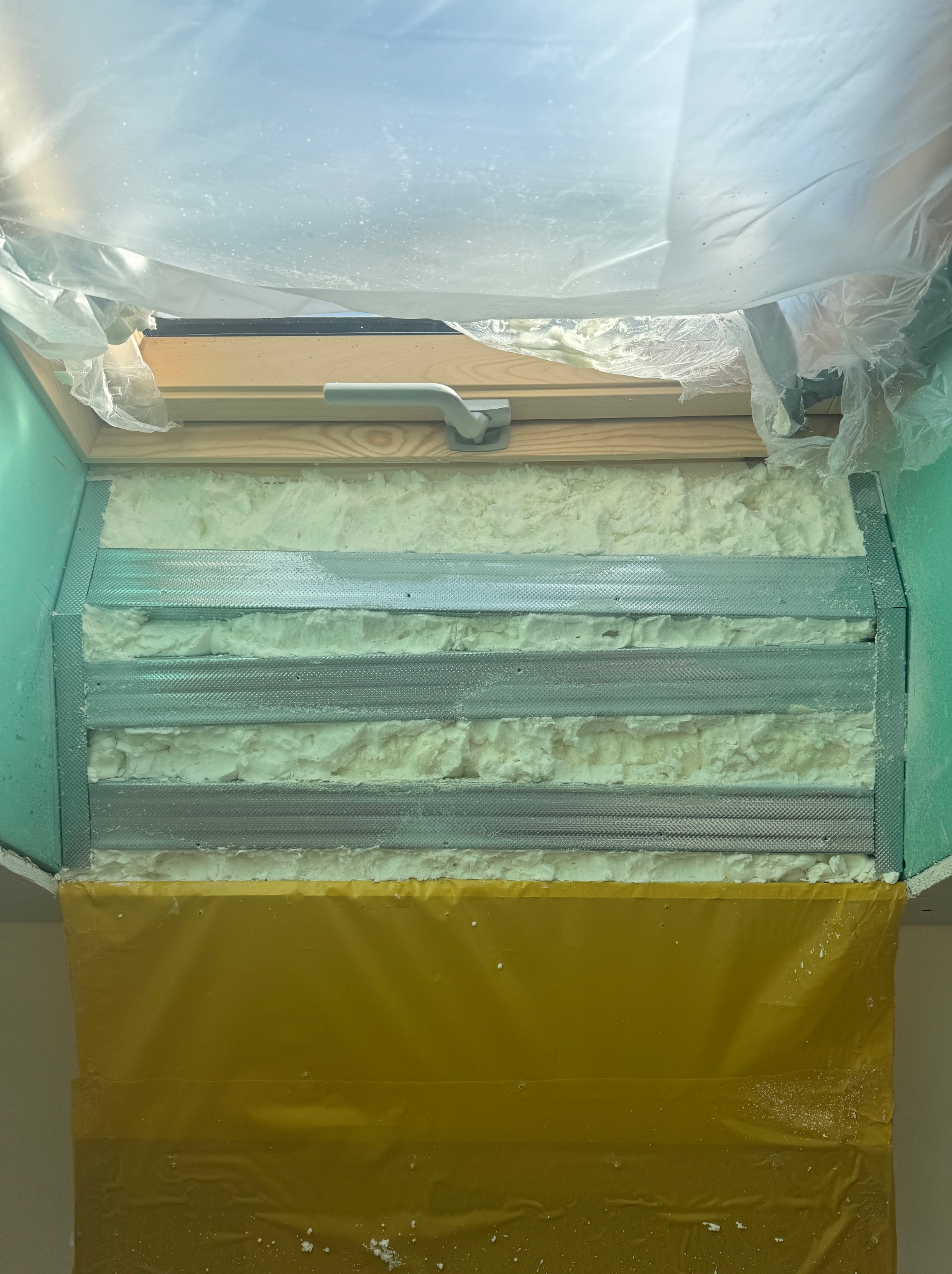 Ocieplanie poddasza pianką PUR wokół okna dachowego, widoczne metalowe profile i zabezpieczająca folia ochronna. Aplikacja pianki w trakcie realizacji, przed wykończeniem.