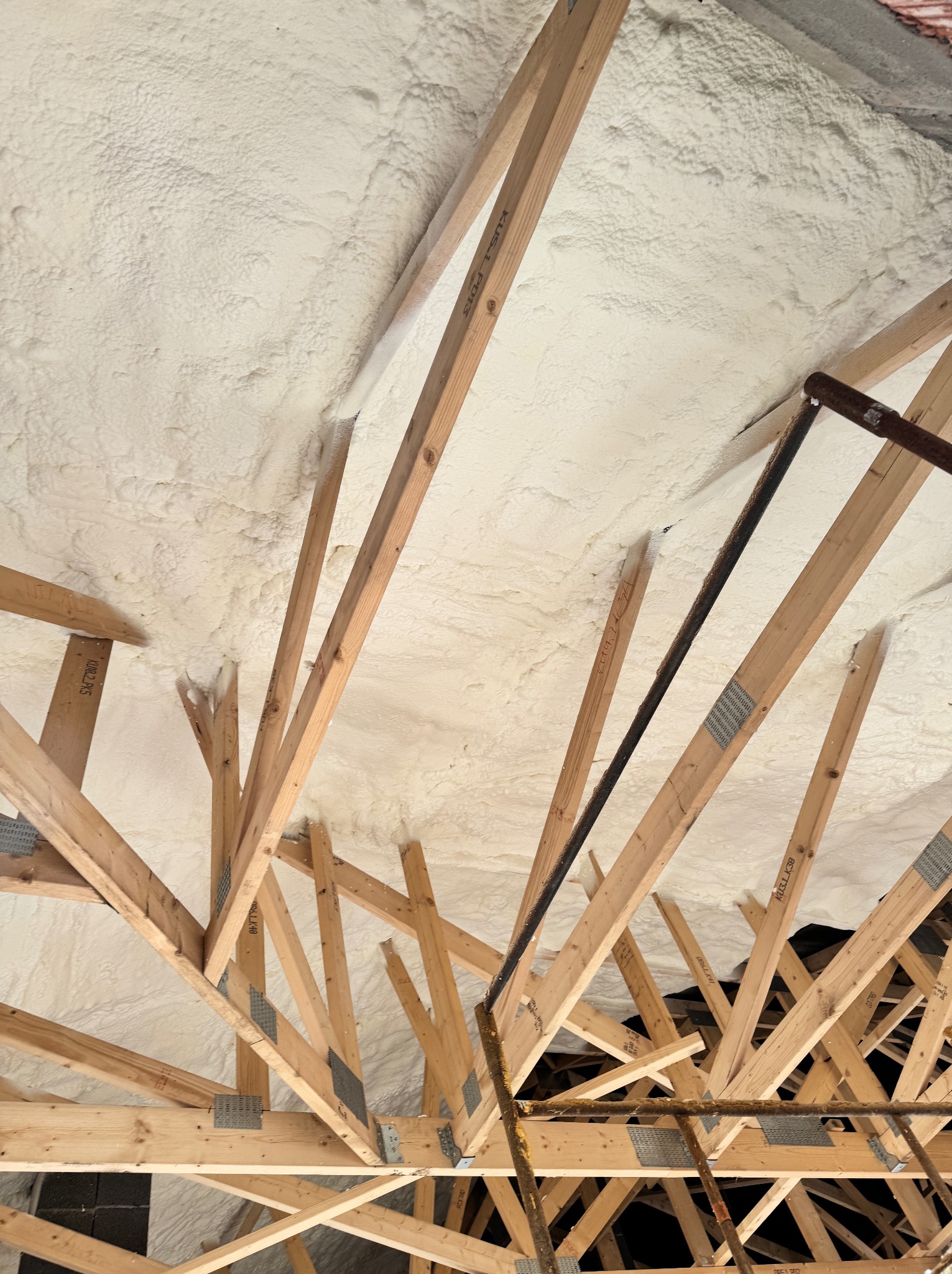Poddasze z drewnianą konstrukcją krokwiową wypełnione białą pianką izolacyjną. Widoczne metalowe łączniki i fragment instalacji. Niski kąt ujęcia.