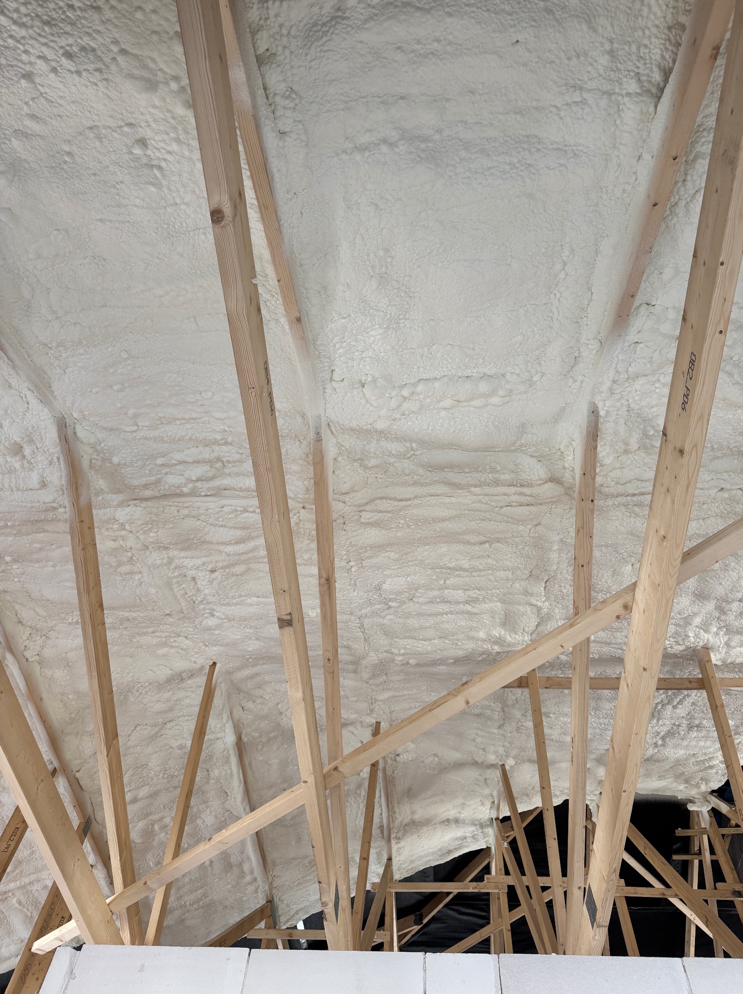 Poddasze z drewnianą konstrukcją i ociepleniem pianką PUR o nieregularnej fakturze, widok z dołu, jasne oświetlenie, fragment ściany z białych bloczków.