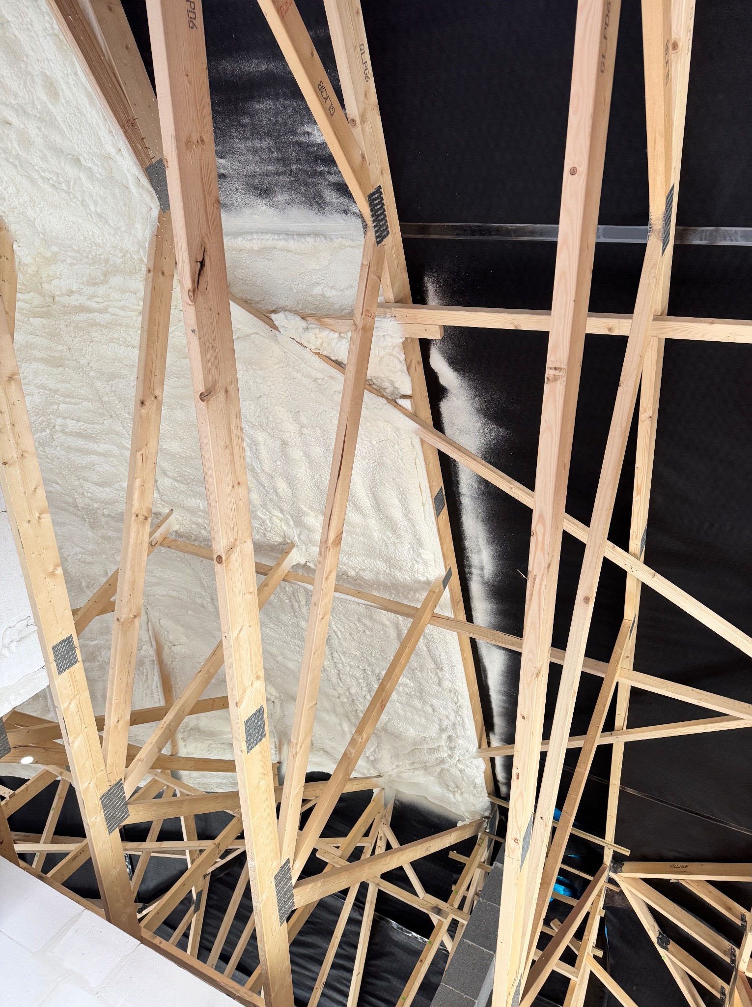 Ocieplenie poddasza pianką PUR, widok od dołu na drewnianą konstrukcję dachu z zaaplikowaną białą pianką izolacyjną i czarną membraną dachową.