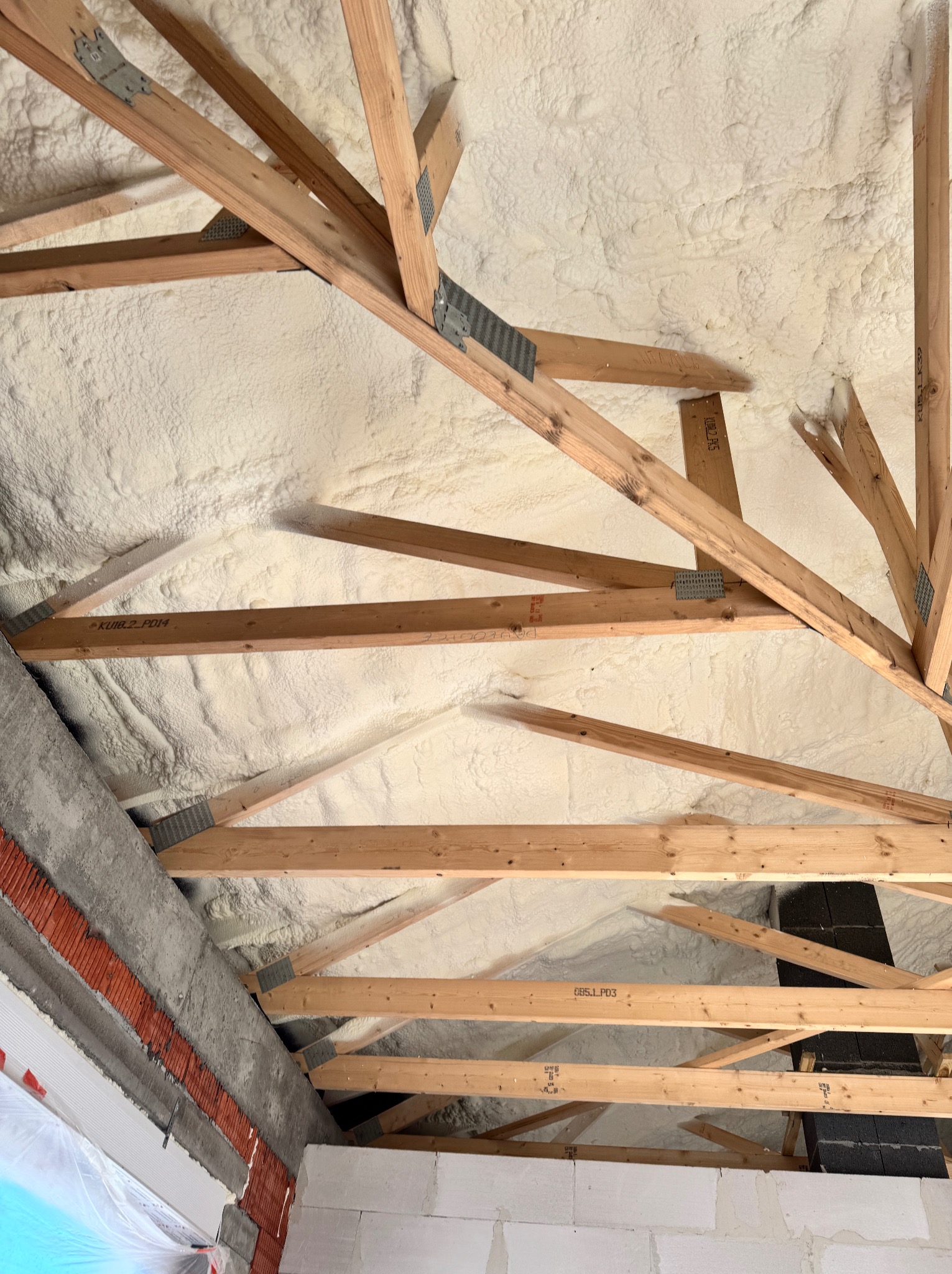 Poddasze po ociepleniu pianką PUR, widoczne drewniane krokwie i szara ściana z ceglanymi elementami. Pianka wypełnia przestrzeń między krokwiami.