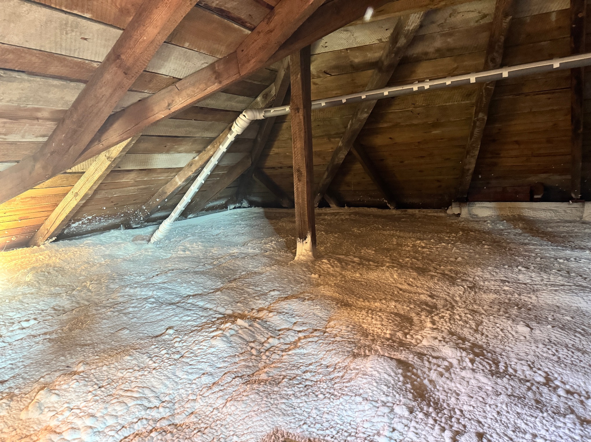 Poddasze po ociepleniu pianką PUR, widoczne drewniane elementy konstrukcyjne i biała, nierówna powierzchnia izolacji. Rury instalacyjne na suficie.