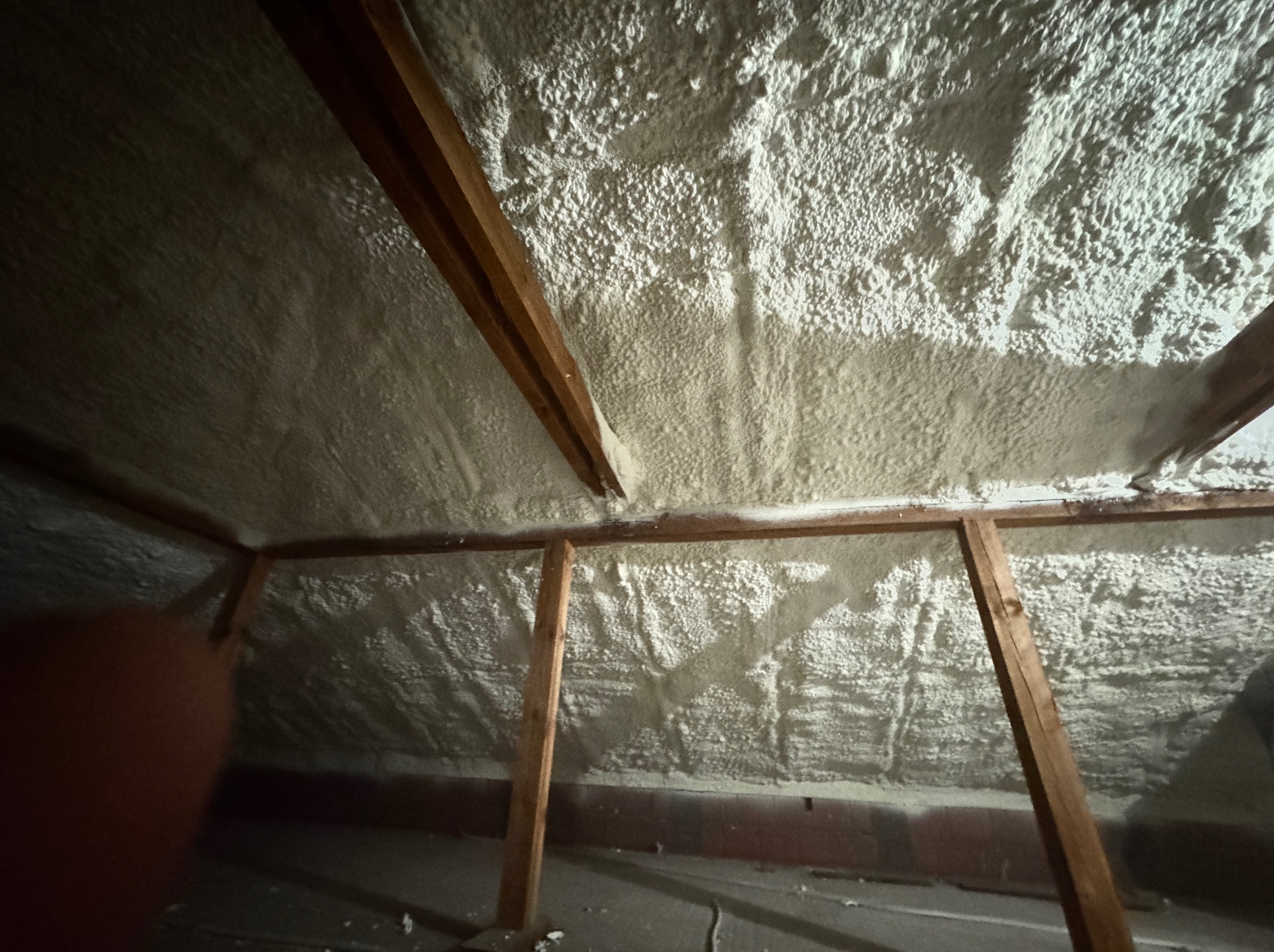 Poddasze z drewnianą konstrukcją pokryte grubą warstwą białej pianki PUR, widoczne nierówności i struktura izolacji. Oświetlenie podkreśla detale piany.