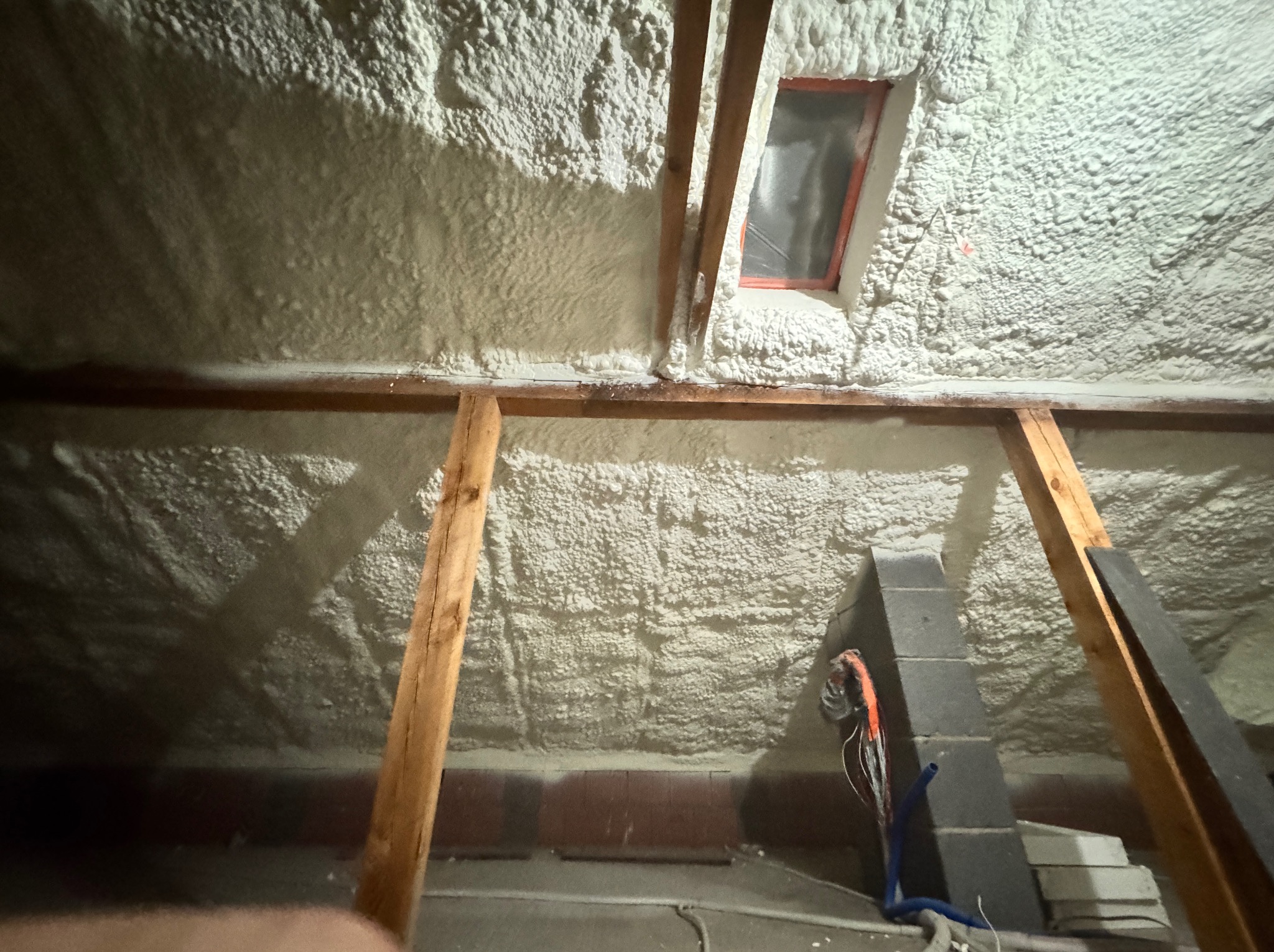 Poddasze po ociepleniu pianką PUR, widoczne drewniane elementy konstrukcyjne, okno dachowe i fragment komina. Szara pianka pokrywa skośne ściany i sufit.