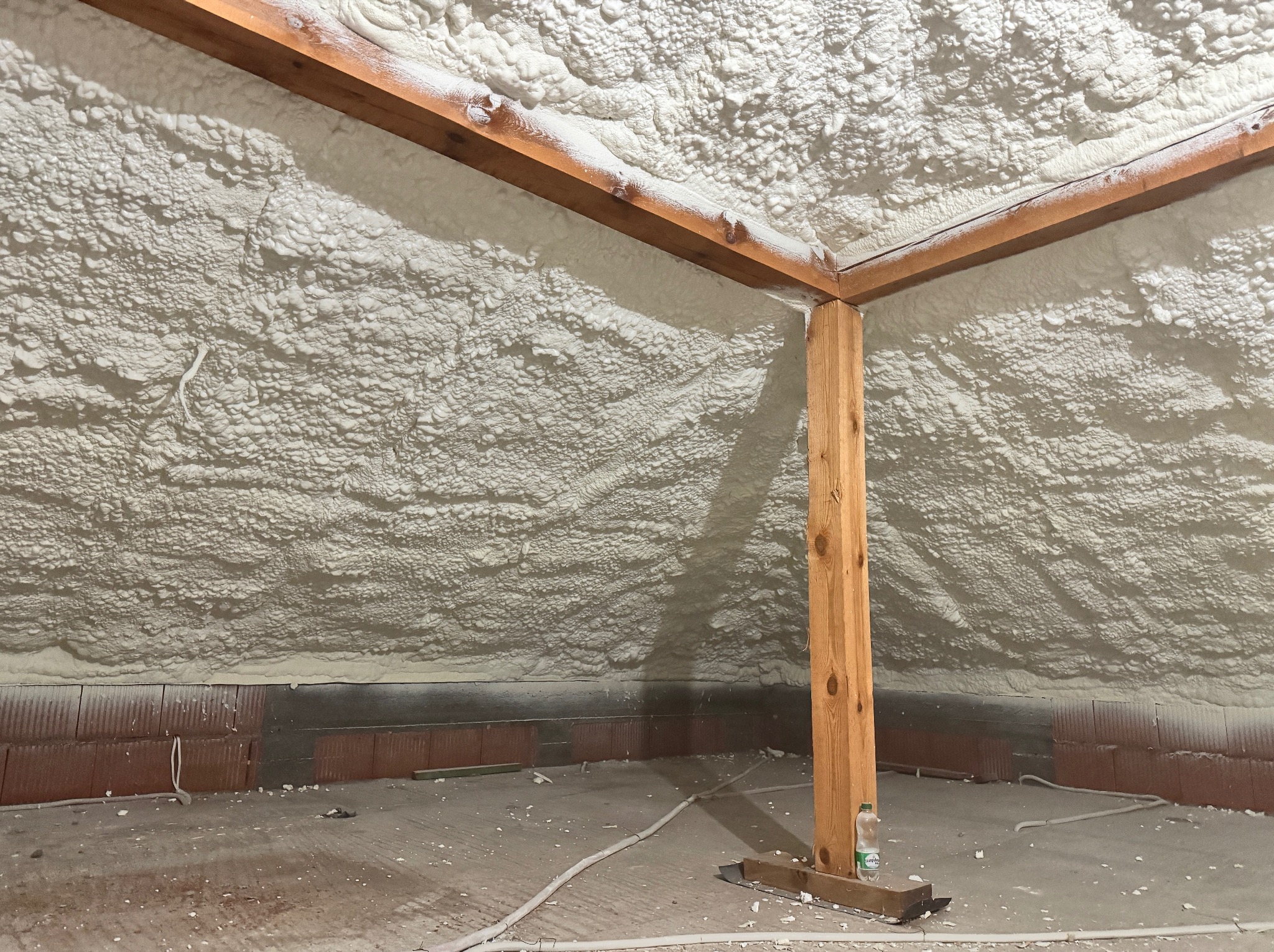 Poddasze po ociepleniu pianką PUR, widoczne drewniane elementy konstrukcyjne i nierównomierna struktura pianki. Fragment ściany z cegły i przewody na podłodze.