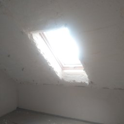 Sigma Pur - Poddasze po ociepleniu pianką PUR, widoczne okno dachowe z intensywnym światłem. Biała pianka pokrywa skosy, sufit i fragment ściany. Szara podłoga w dolnej części kadru.