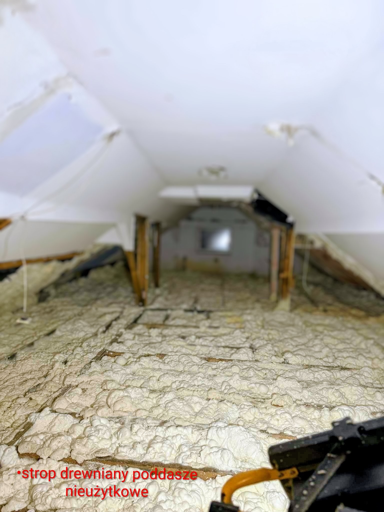 Poddasze z drewnianym stropem po ociepleniu pianką PUR, widoczne krokwie i biała, nierówna powierzchnia izolacji. Niewykończone wnętrze.