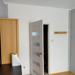 Mateusz - Otwarta, nowoczesna, biała drzwi wewnętrzne z przeszkleniami, zamontowana w odnowionym pokoju z szaro-białymi ścianami i drewnianą półką. Widoczny fragment szafy.