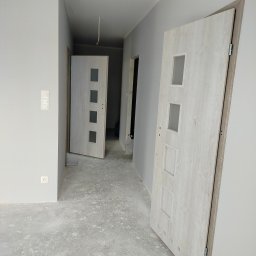 Mateusz - Wnętrze w szarościach z dwiema białymi, częściowo zamontowanymi drzwiami z czterema kwadratowymi przeszkleniami. Podłoga w trakcie prac wykończeniowych.