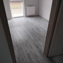 Mateusz - Pokój po remoncie: szare panele podłogowe imitujące drewno, białe ściany, okno z widokiem na zewnątrz i grzejnik. Widok zza otwartych drzwi, częściowo widoczna ościeżnica.