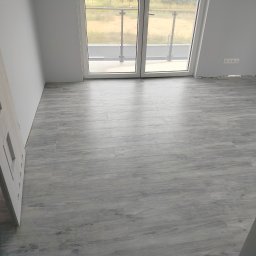 Mateusz - Jasnoszara podłoga z płytek imitujących drewno w pokoju z białymi ścianami, widoczne drzwi i balkon z przeszkloną balustradą, gniazdka elektryczne przy podłodze.