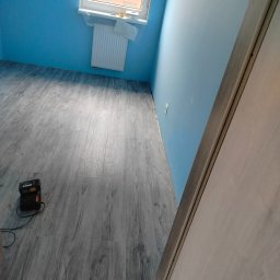 Mateusz - Pokój po remoncie: świeżo położona podłoga z paneli w odcieniach szarości, błękitne ściany i białe drzwi. Widoczny grzejnik pod oknem i ładowarka do narzędzi.