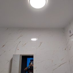 Mateusz - Wykończona łazienka z białymi płytkami imitującymi marmur, oświetlona okrągłą lampą sufitową. Widoczne lustro i instalacja. Osoba robi zdjęcie w lustrze.