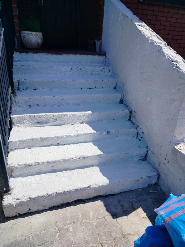 Białe, świeżo pomalowane betonowe schody wejściowe z czarną balustradą i rośliną doniczkową w tle. Widoczne ślady niedokładnego malowania.