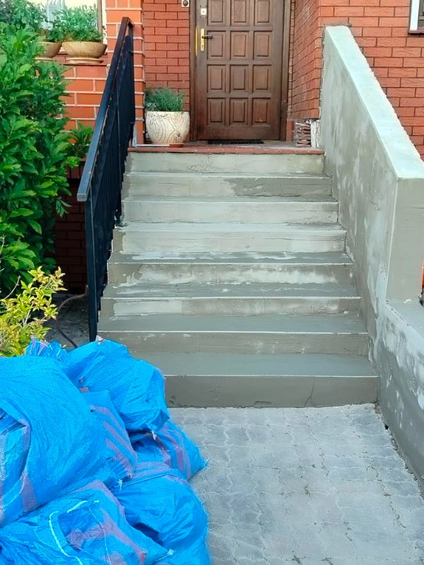 Odnowione betonowe schody wejściowe do domu z cegły, z czarną balustradą i workami z materiałem budowlanym u dołu. Widoczne ślady świeżych prac remontowych.