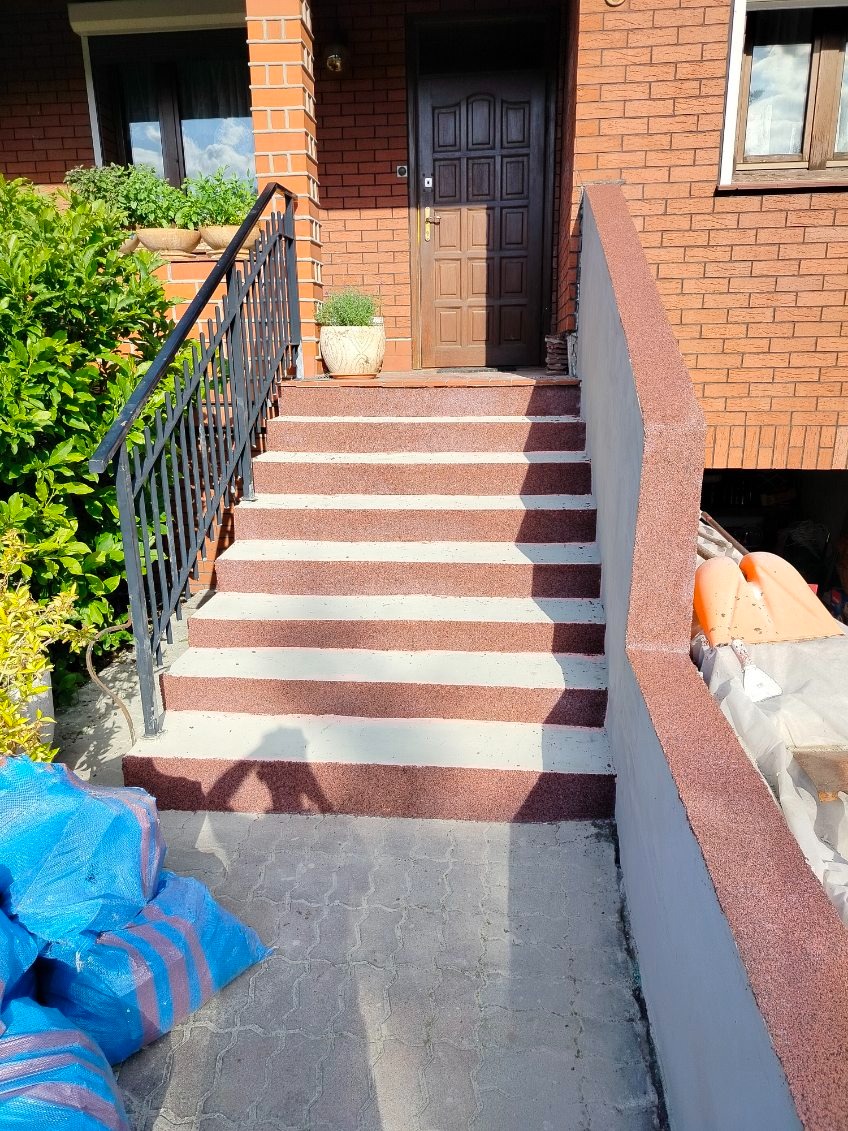 Odnowione schody wejściowe do domu z brązową elewacją, wykończone żywicą w kolorze terakoty i bieli. Balustrada z czarnego metalu. Widoczne worki z materiałami budowlanymi.