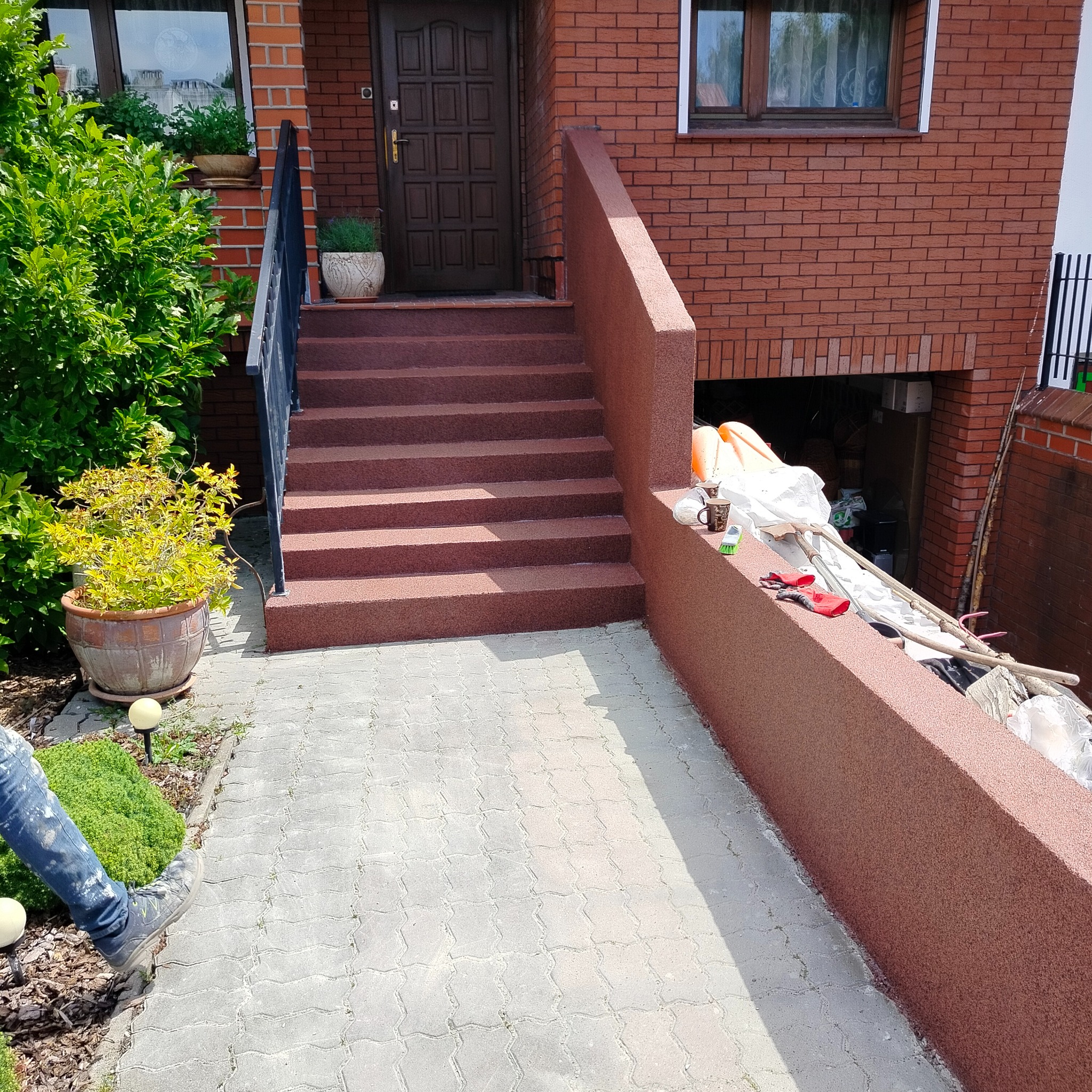 Odnowione, brązowe schody wejściowe do domu z cegły. Świeżo pomalowane elementy architektoniczne, doniczki z roślinami. Widoczne narzędzia i akcesoria malarskie.