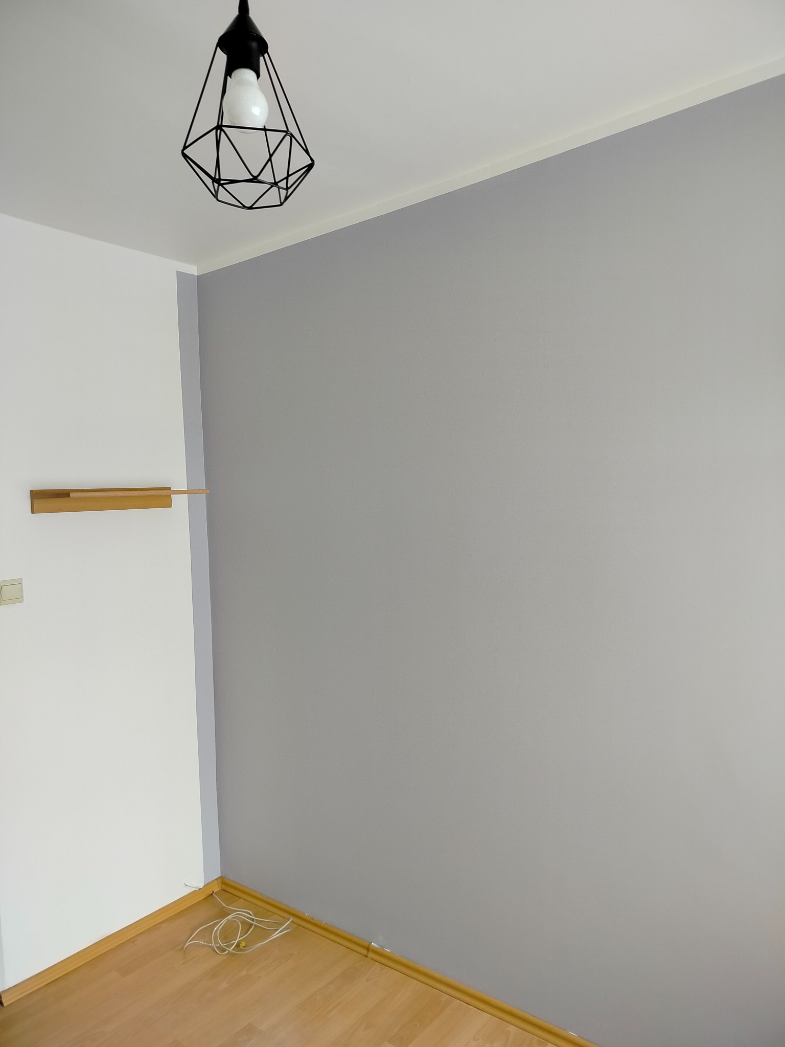 Pomalowany na szaro pokój z drewnianą półką na białej ścianie i nowoczesną lampą. Widoczne kable na podłodze. Świeżo odmalowane ściany w pokoju.