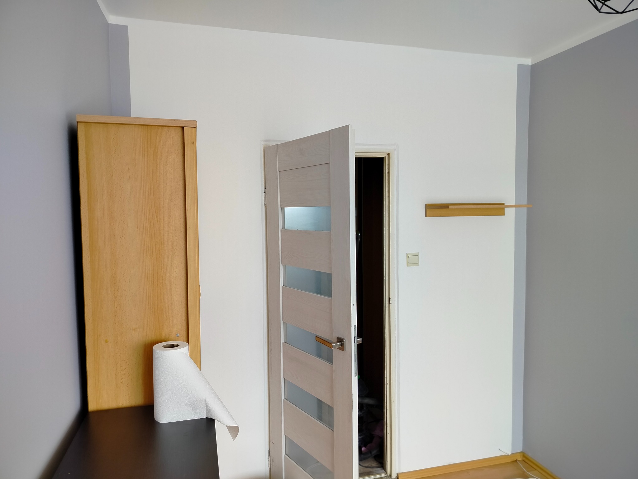 Otwarta, nowoczesna, biała drzwi wewnętrzne z przeszkleniami, zamontowana w odnowionym pokoju z szaro-białymi ścianami i drewnianą półką. Widoczny fragment szafy.