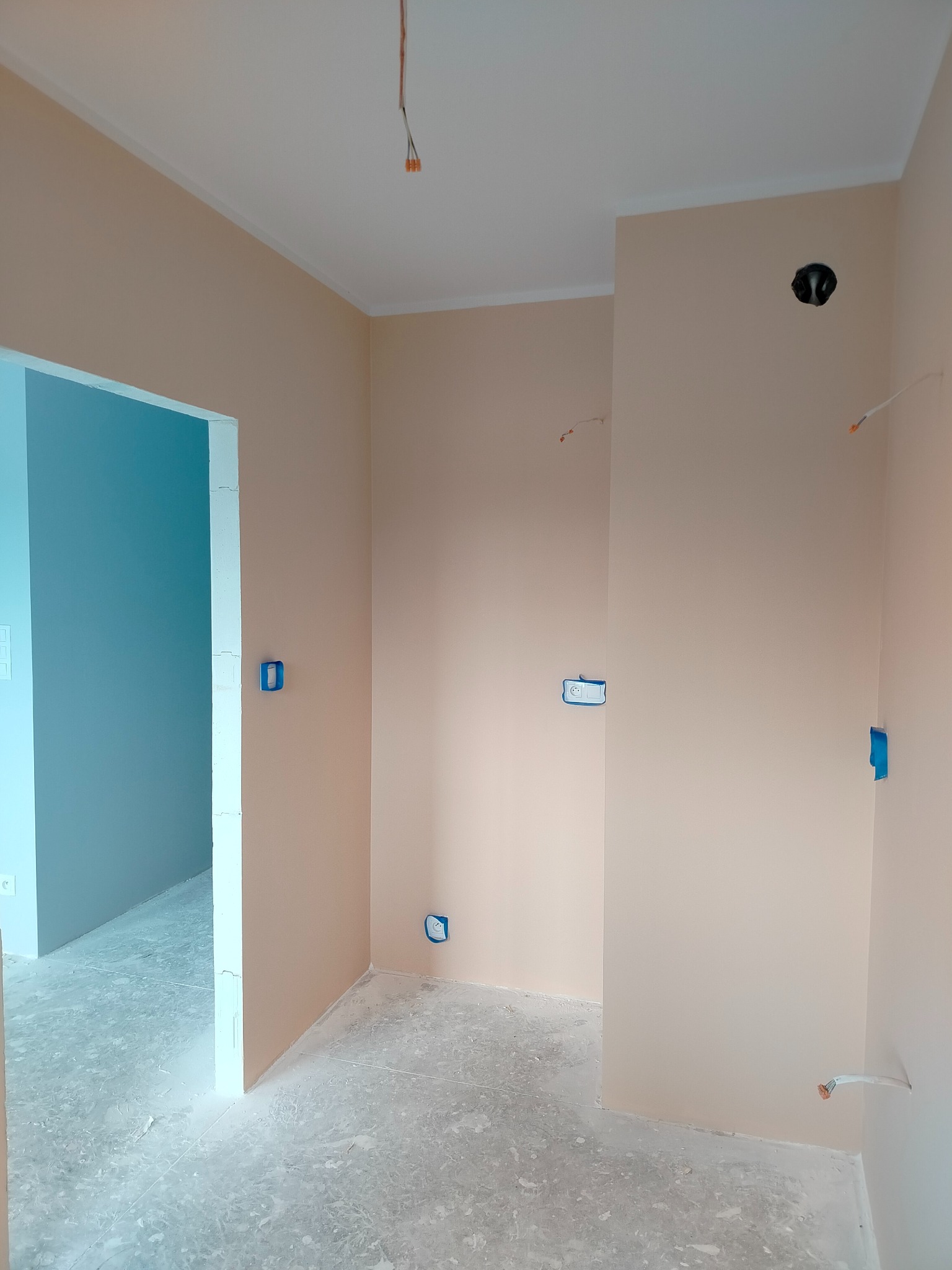 Pokój po malowaniu ścian na beżowo, z widocznymi puszkami elektrycznymi zabezpieczonymi taśmą malarską i otwartym przejściem do innego pomieszczenia. Wykończenie w trakcie.