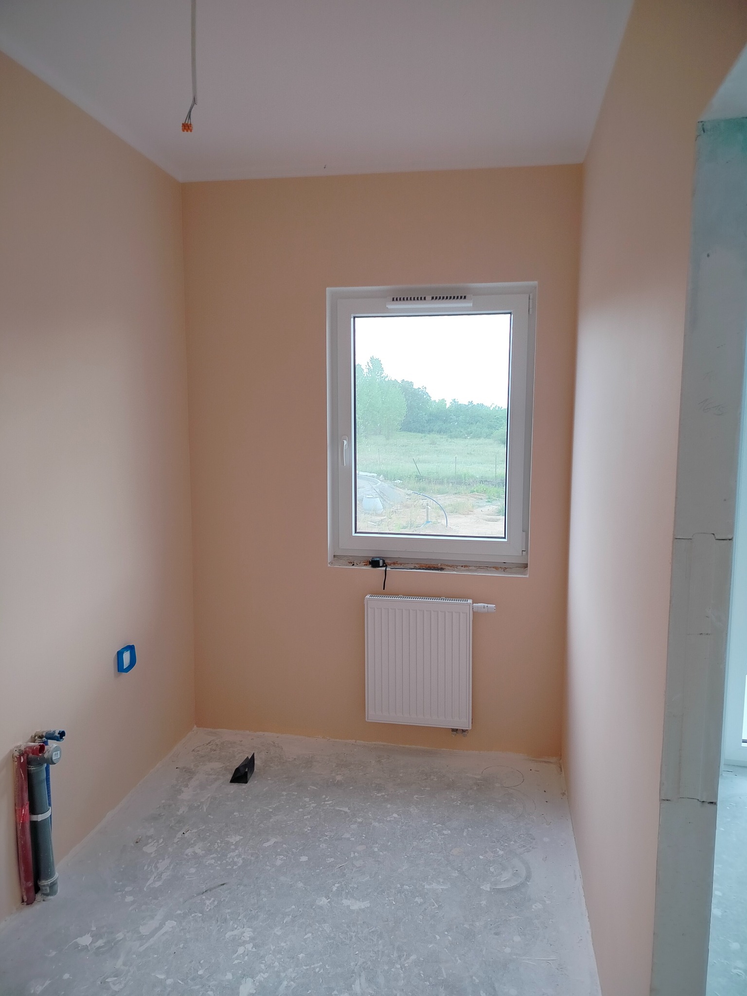Pomieszczenie po malowaniu na brzoskwiniowo z oknem na zewnątrz. Widoczne rury, grzejnik i surowa podłoga. Przygotowane do dalszych prac wykończeniowych.
