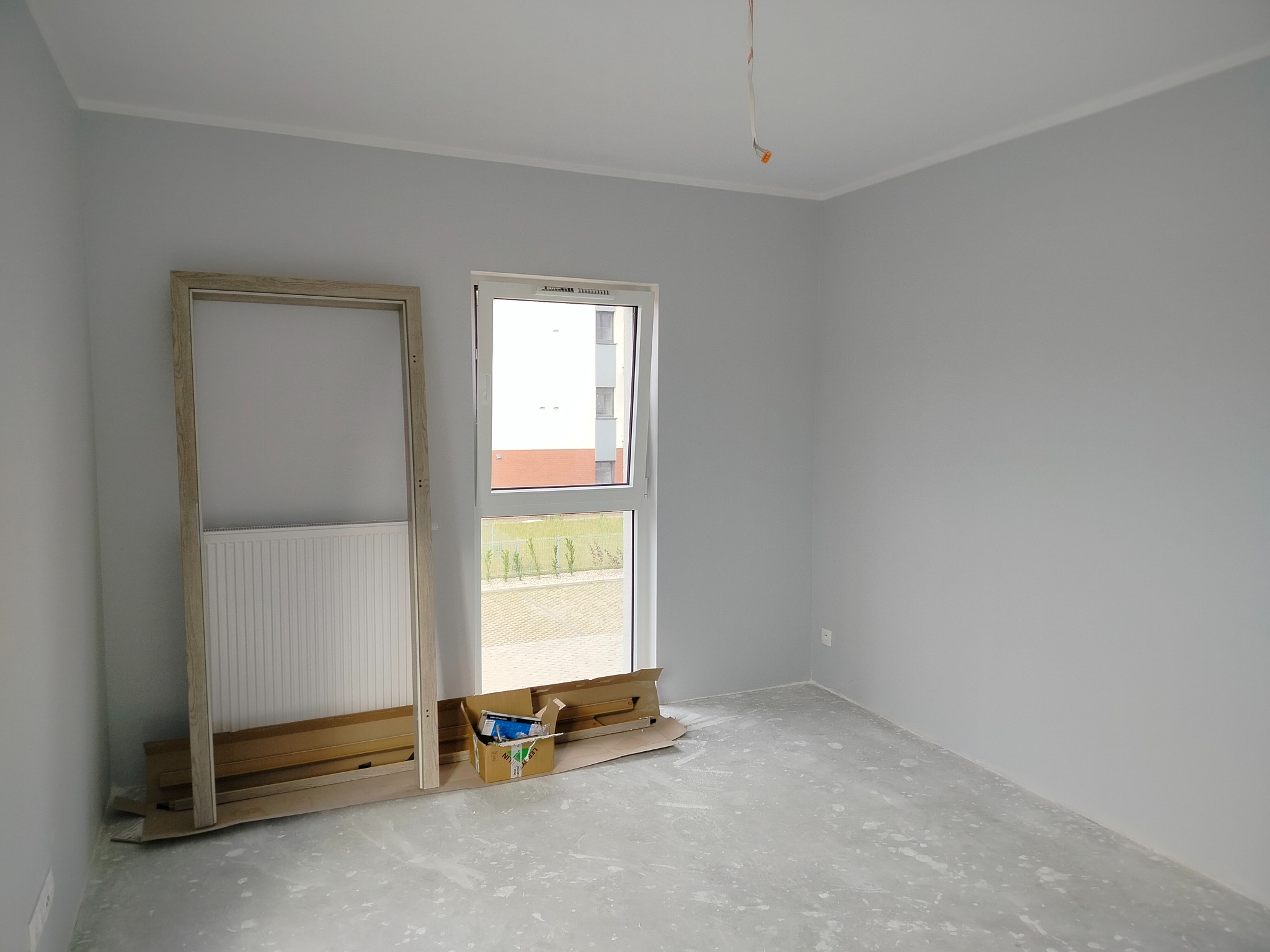 Przygotowanie do montażu drzwi w Bydgoszczy: ościeżnica oparta o ścianę, okno z widokiem na zewnątrz, szare ściany i podłoga w trakcie wykończenia.