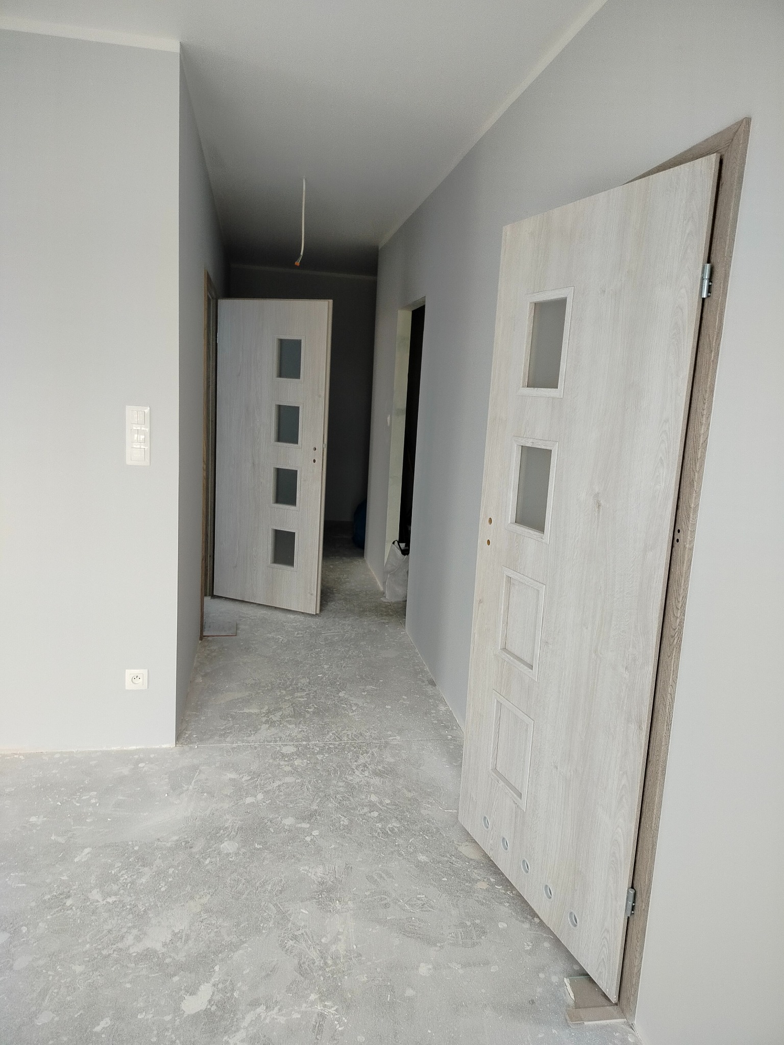 Wnętrze w szarościach z dwiema białymi, częściowo zamontowanymi drzwiami z czterema kwadratowymi przeszkleniami. Podłoga w trakcie prac wykończeniowych.