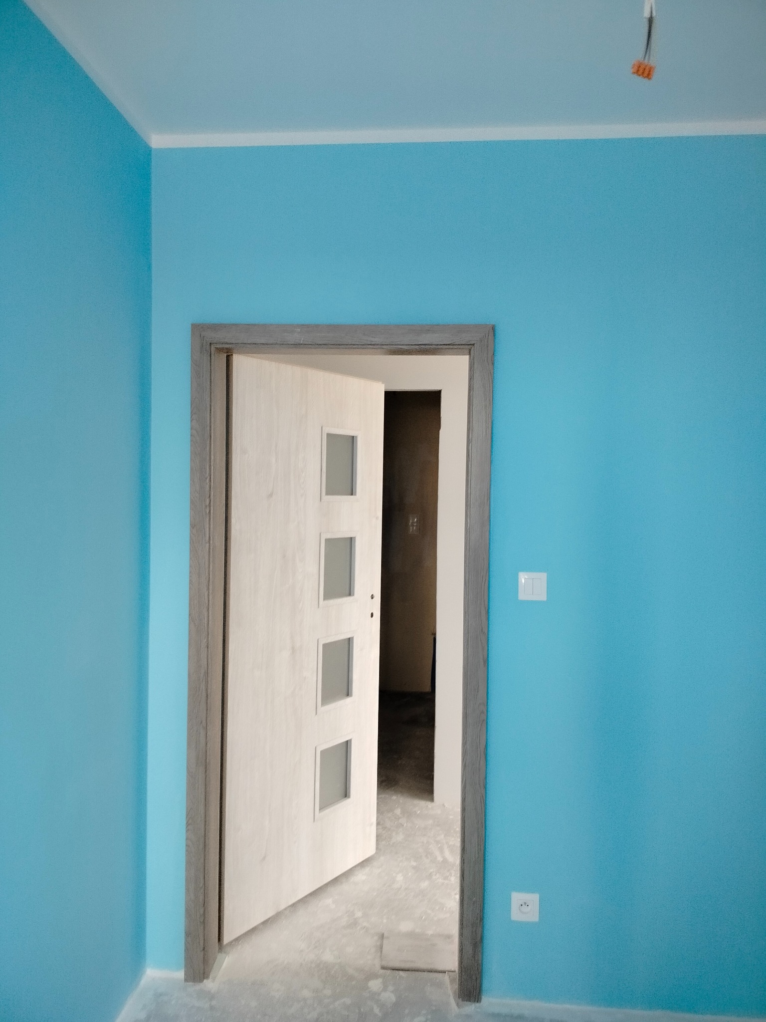 Świeżo pomalowane na błękitno ściany z zamontowanymi drzwiami z czterema szybkami i szarą futryną. Widoczny fragment gniazdka elektrycznego i włącznika.