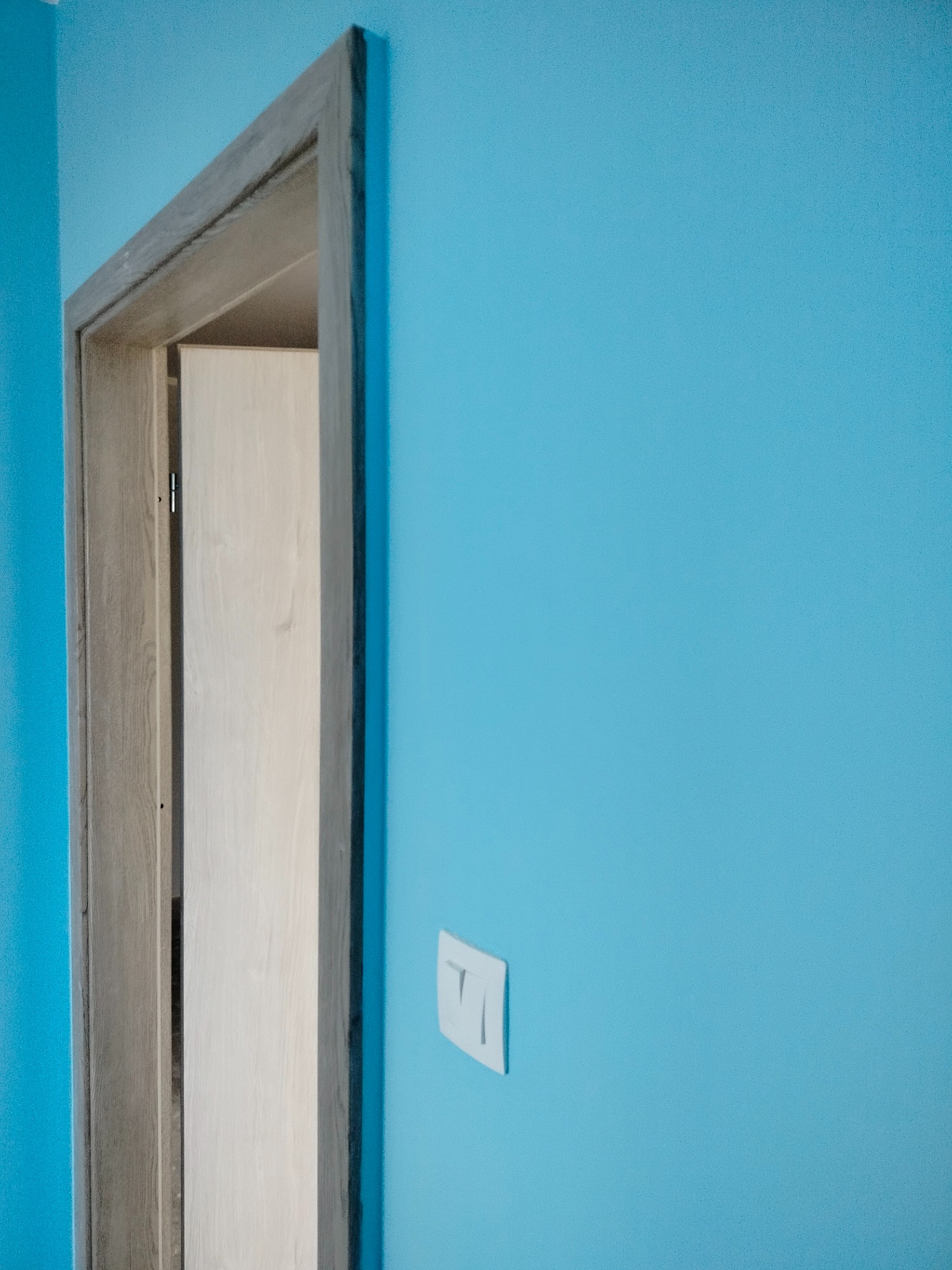 Turkusowa ściana z zamontowaną ościeżnicą drzwiową w surowym stanie i włącznikiem światła. Widok na wnętrze.