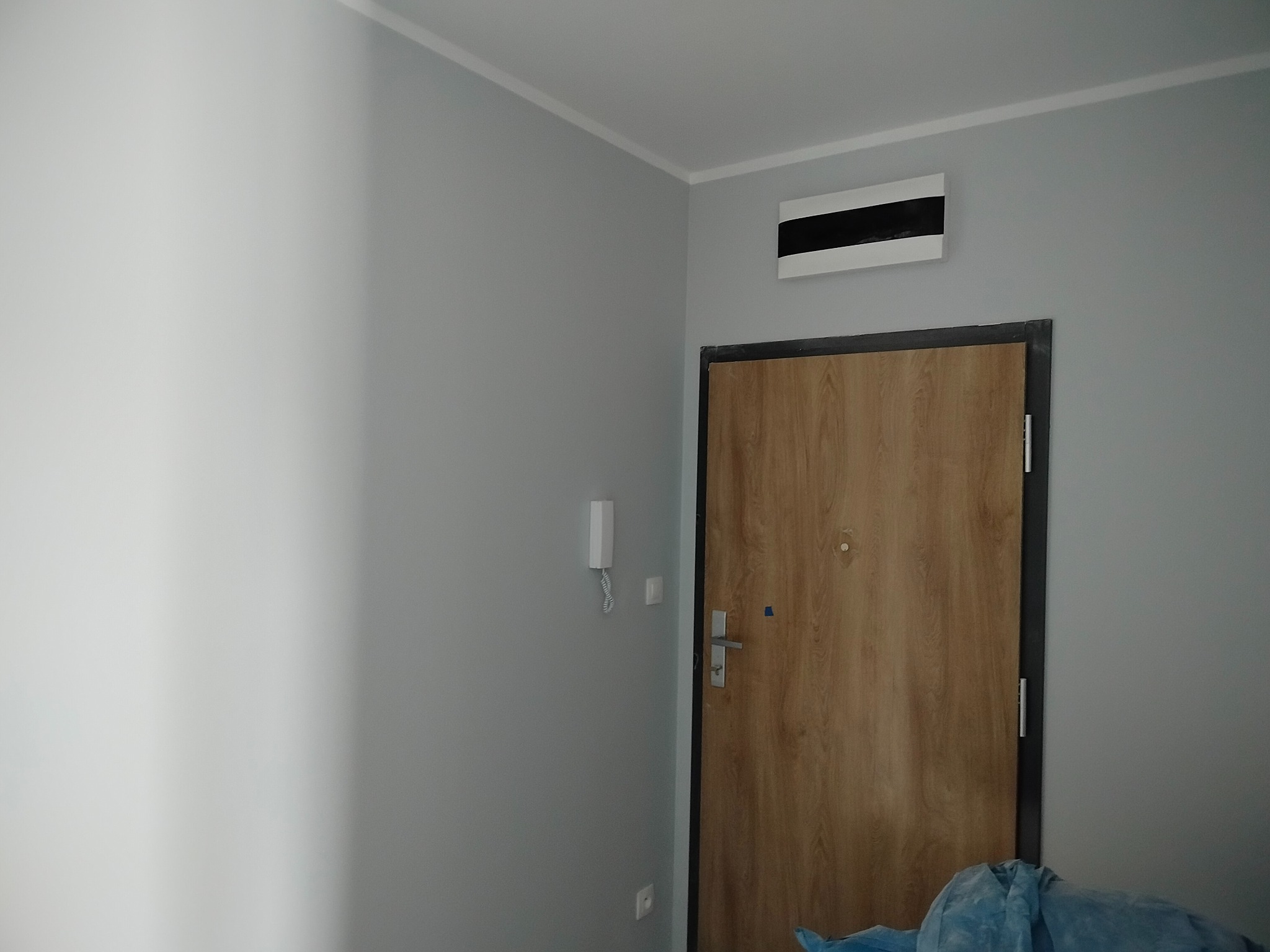 Szare ściany po malowaniu w przedpokoju, widoczna drewniana ościeżnica i drzwi. Na ścianie domofon i skrzynka elektryczna. U dołu fragment zabezpieczenia podłogi.