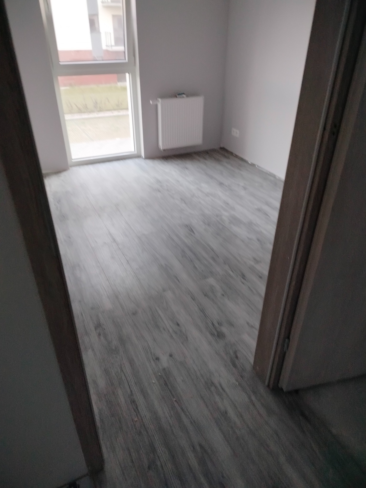Pokój po remoncie: szare panele podłogowe imitujące drewno, białe ściany, okno z widokiem na zewnątrz i grzejnik. Widok zza otwartych drzwi, częściowo widoczna ościeżnica.