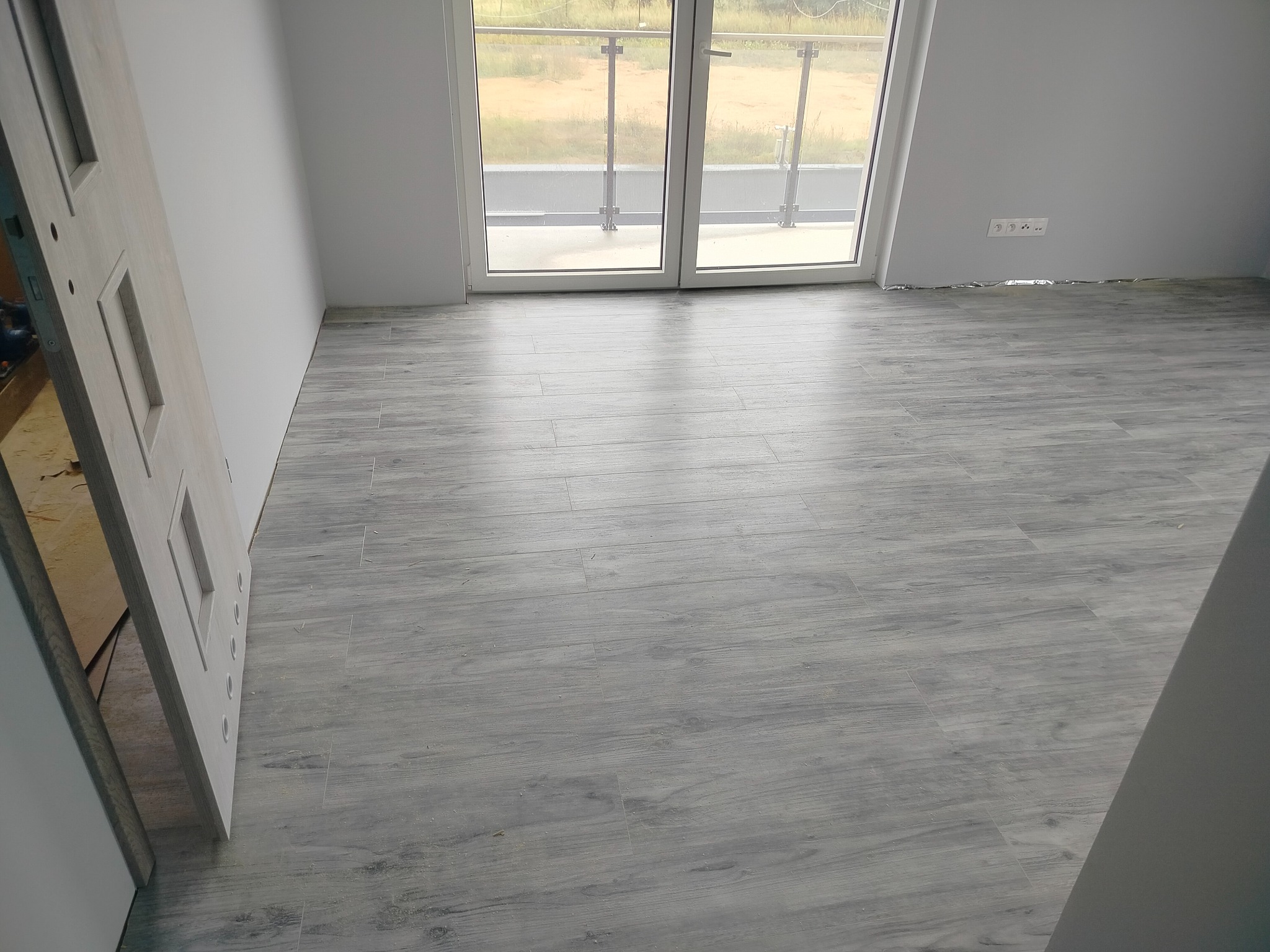 Jasnoszara podłoga z płytek imitujących drewno w pokoju z białymi ścianami, widoczne drzwi i balkon z przeszkloną balustradą, gniazdka elektryczne przy podłodze.