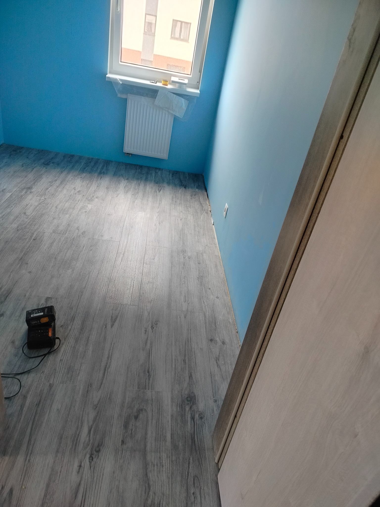 Pokój po remoncie: świeżo położona podłoga z paneli w odcieniach szarości, błękitne ściany i białe drzwi. Widoczny grzejnik pod oknem i ładowarka do narzędzi.