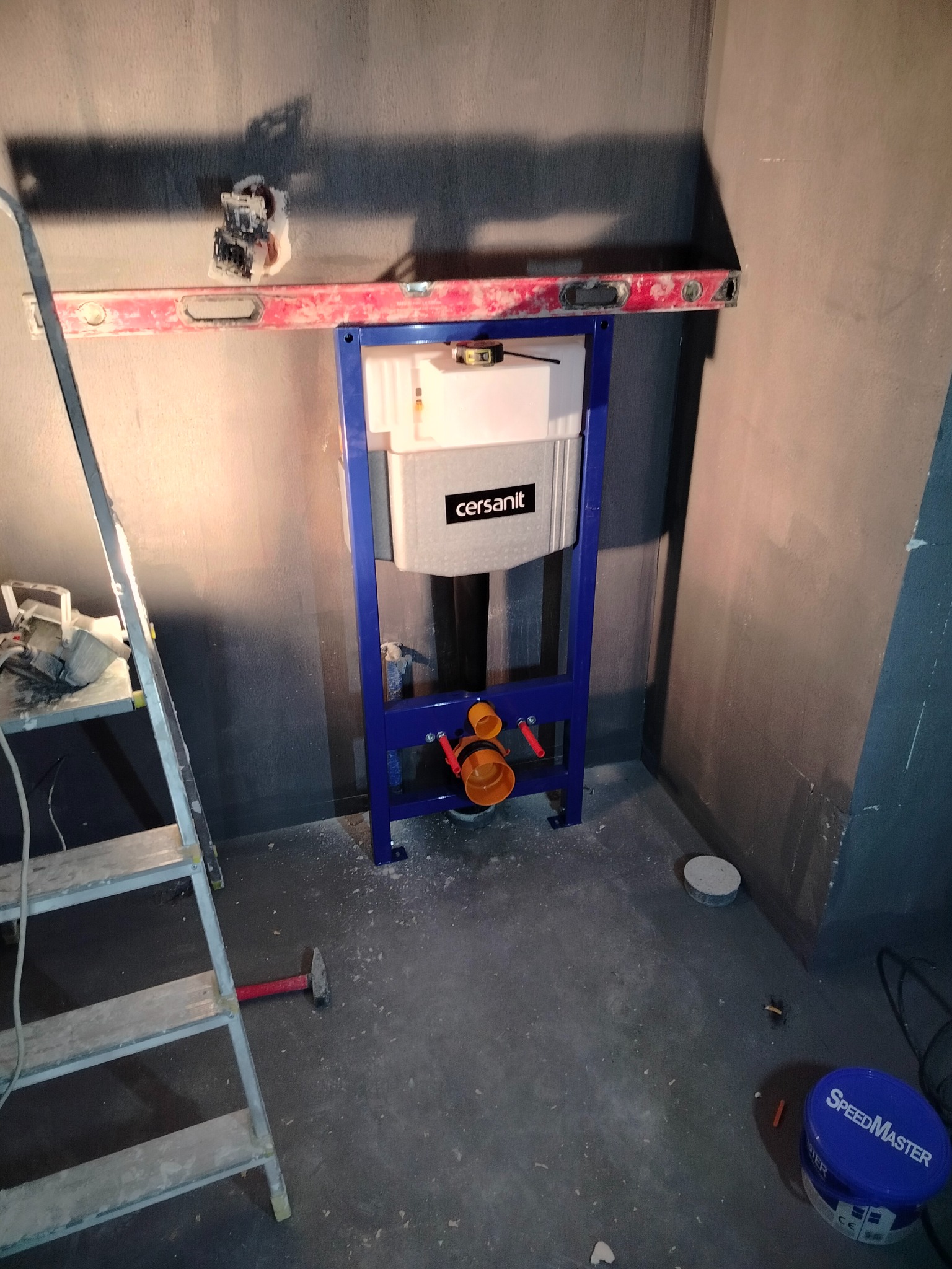 Stelaż podtynkowy Cersanit zamontowany w surowej łazience, poziomowany z użyciem poziomicy. Widoczna drabina i narzędzia. Przygotowanie do wykończenia łazienki.