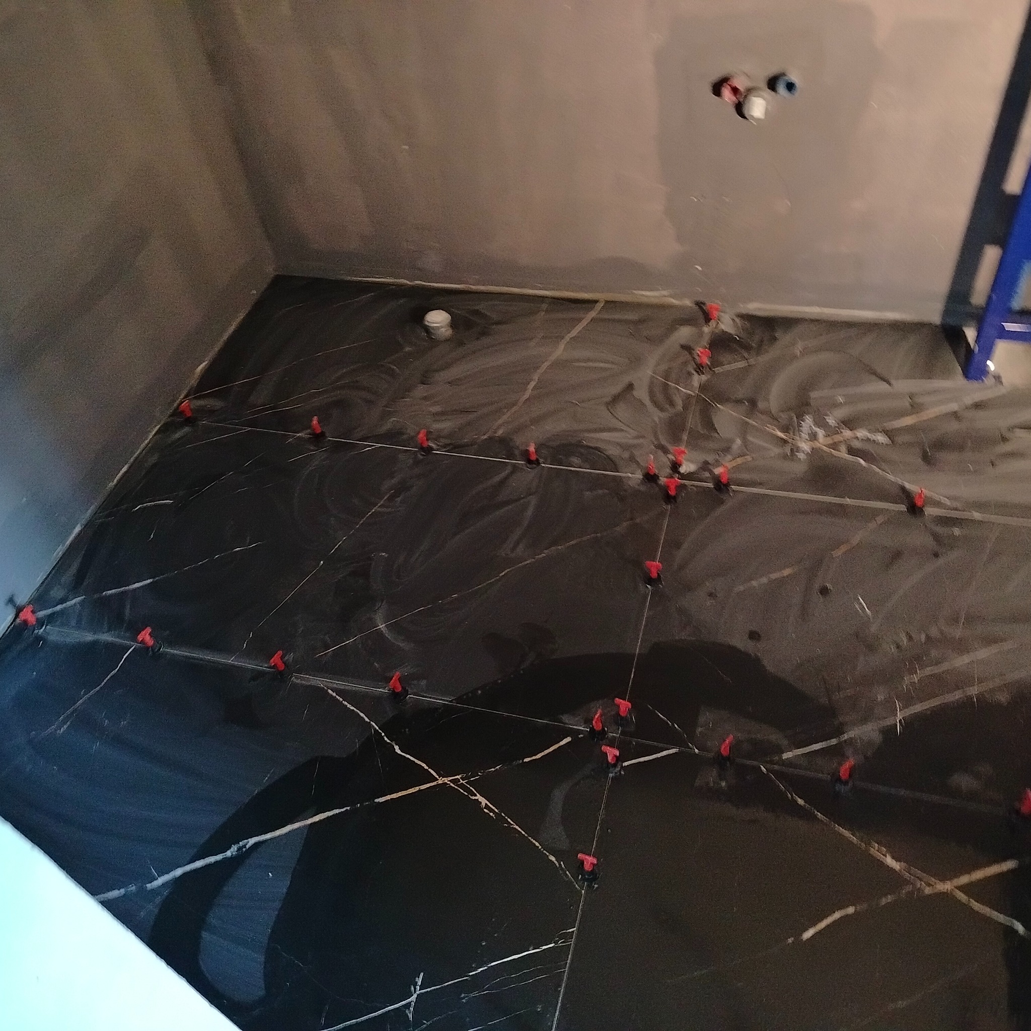 Czarne płytki z białym wzorem marmuru, wyrównane za pomocą czerwonych klipsów w trakcie wykańczania łazienki; widoczne przyłącza wodne i drabina.