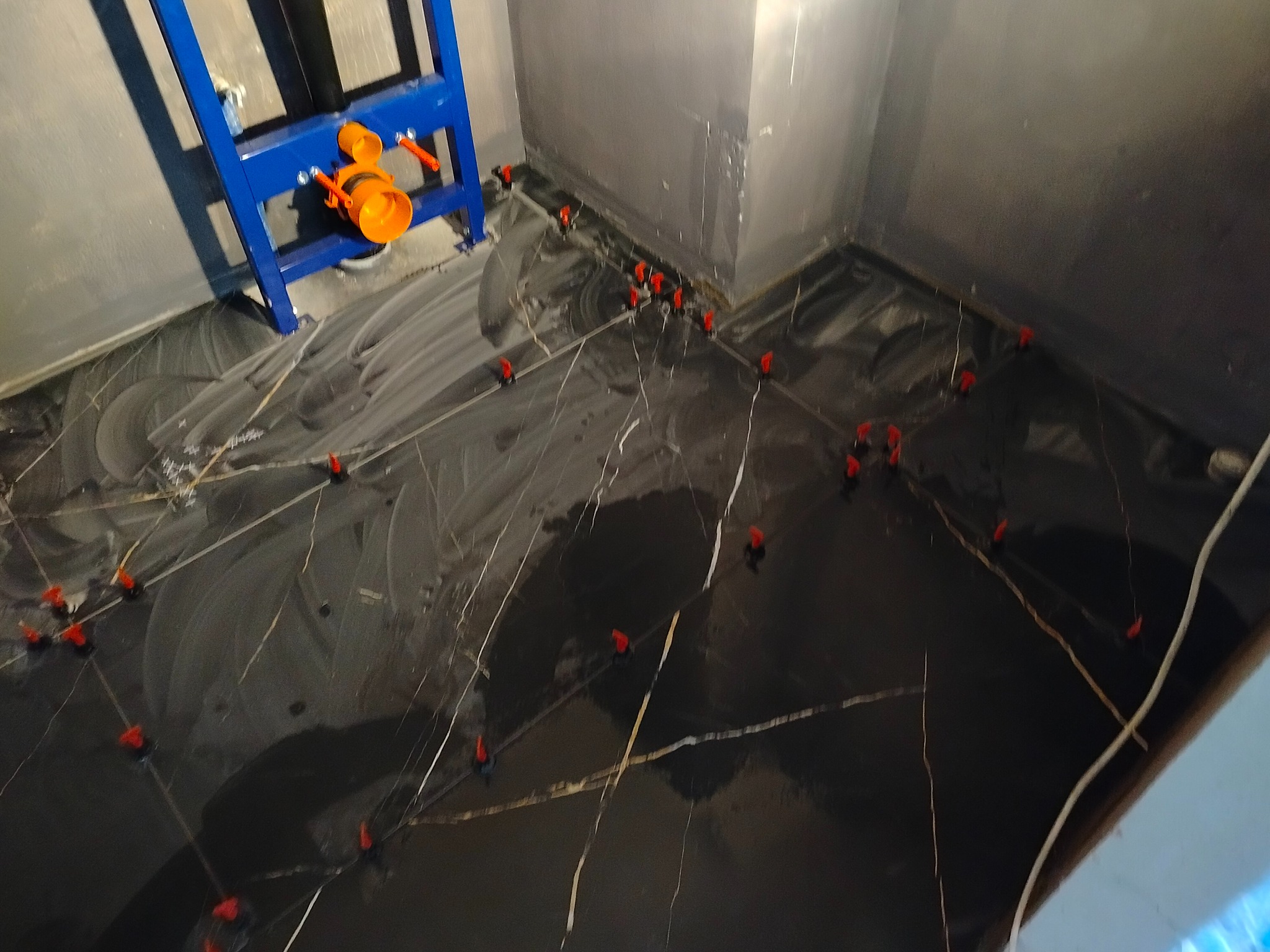 Ciemne płytki podłogowe w łazience, ułożone z użyciem czerwonych klinów poziomujących. W tle widoczny stelaż podtynkowy WC w kolorze niebieskim.