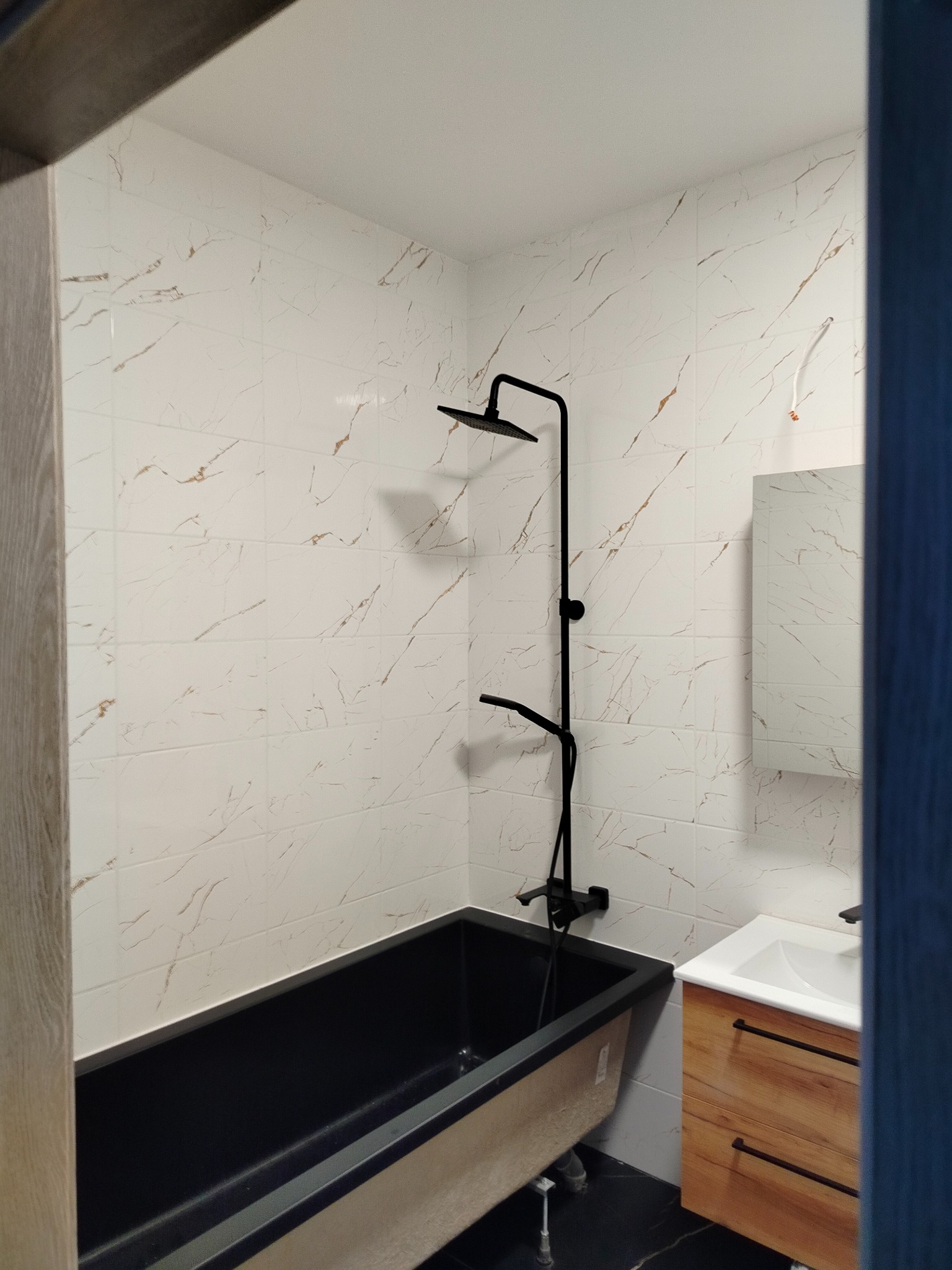 Nowoczesna łazienka po remoncie: czarna wanna na tle białych płytek imitujących marmur, czarna armatura i drewniana szafka pod umywalką. Widok zza framugi.