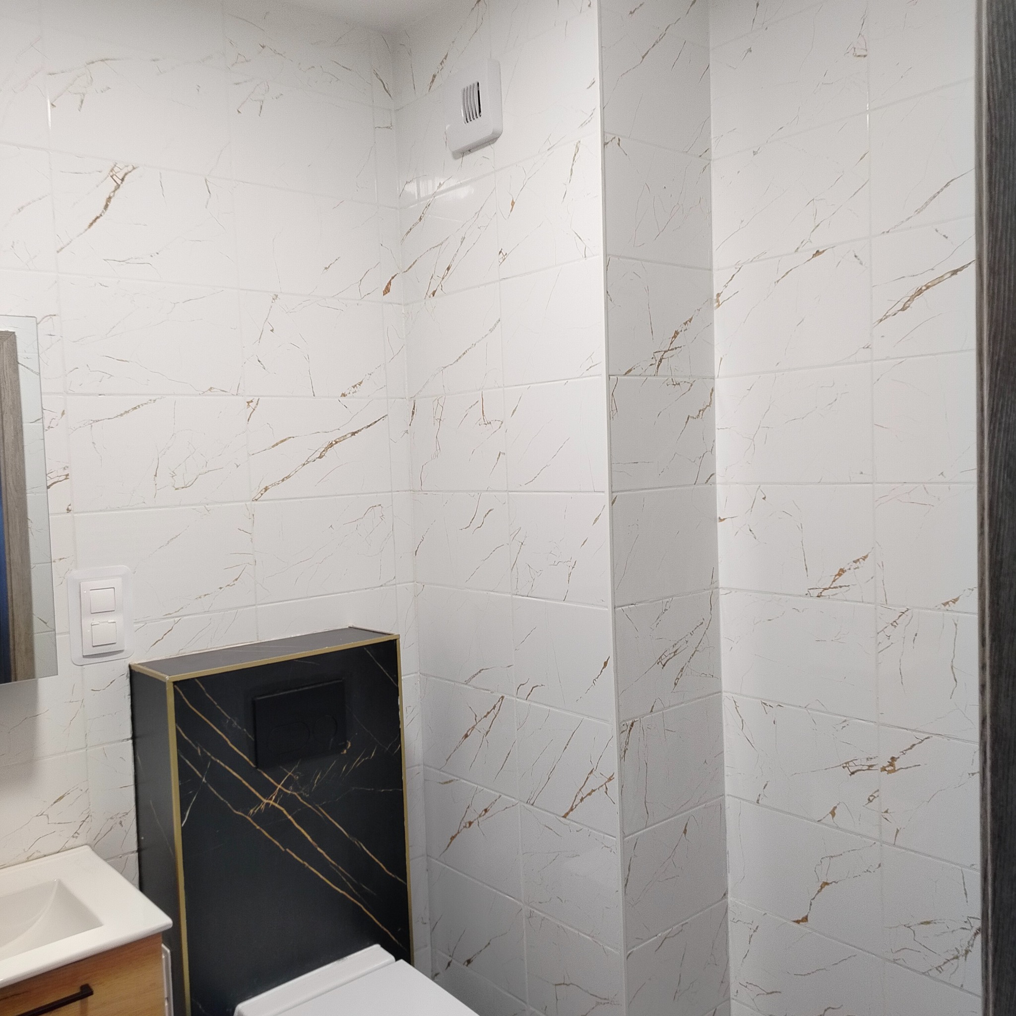 Wykończona łazienka z białymi płytkami imitującymi marmur ze złotymi żyłami, zabudowa WC w czarnym marmurze ze złotymi akcentami i biała armatura.