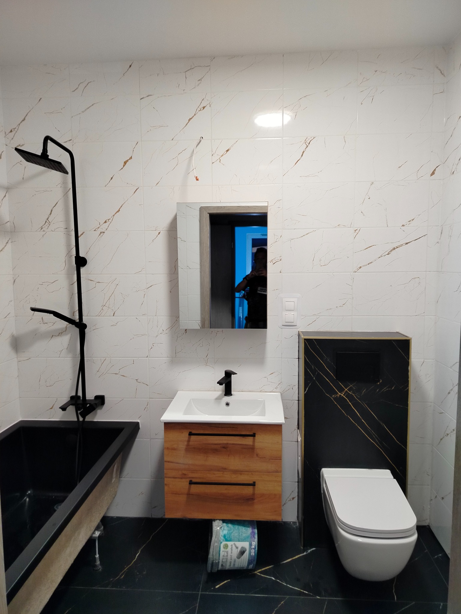 Nowoczesna łazienka z czarną armaturą, wanną i płytkami imitującymi marmur. Umywalka z drewnianą szafką i toaleta w minimalistycznym stylu.